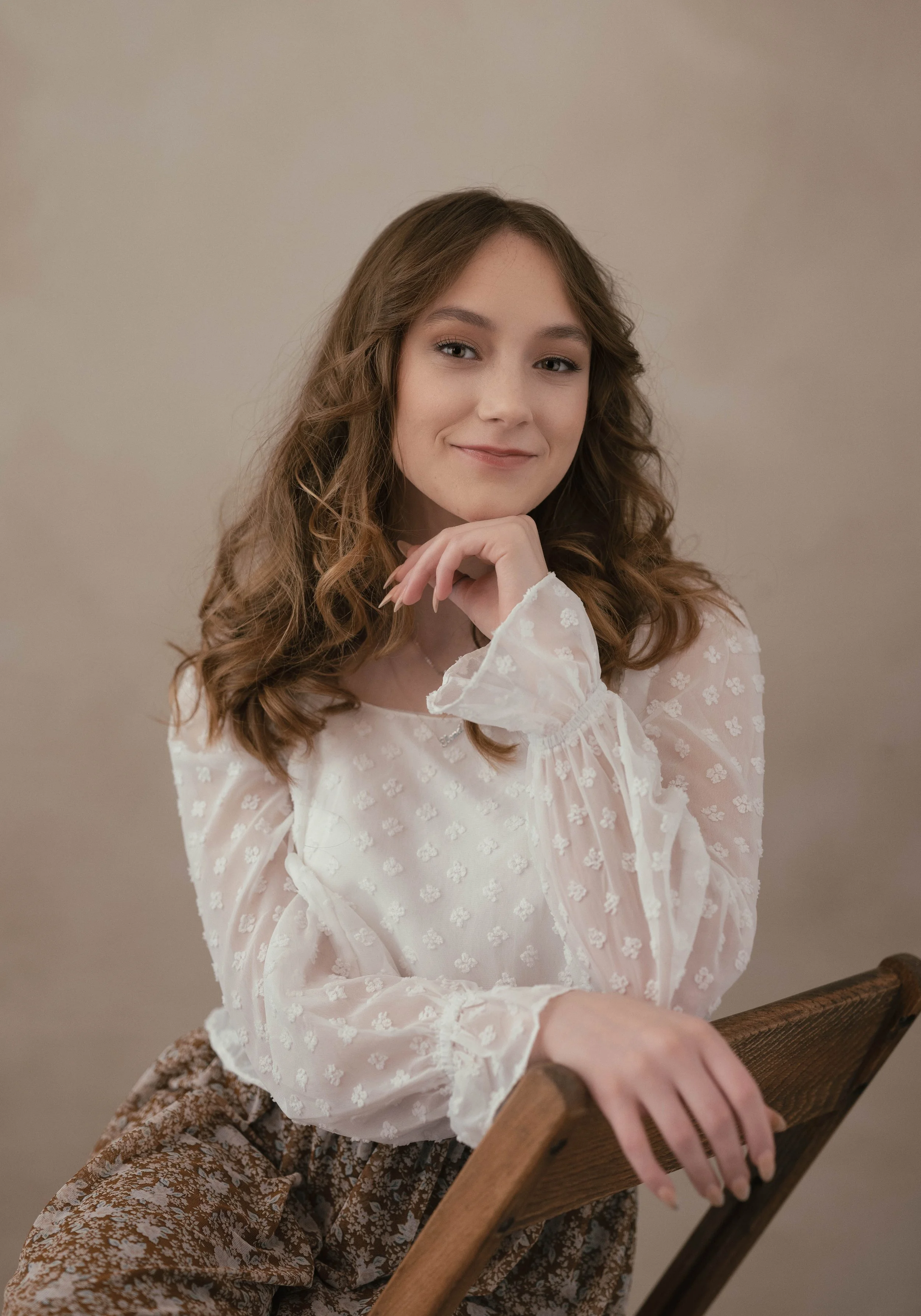 A young woman with wavy brown hair smiling gently, sitting on a wooden chair, wearing a sheer white blouse with floral embroidery and patterned brown pants, against a plain beige wall.