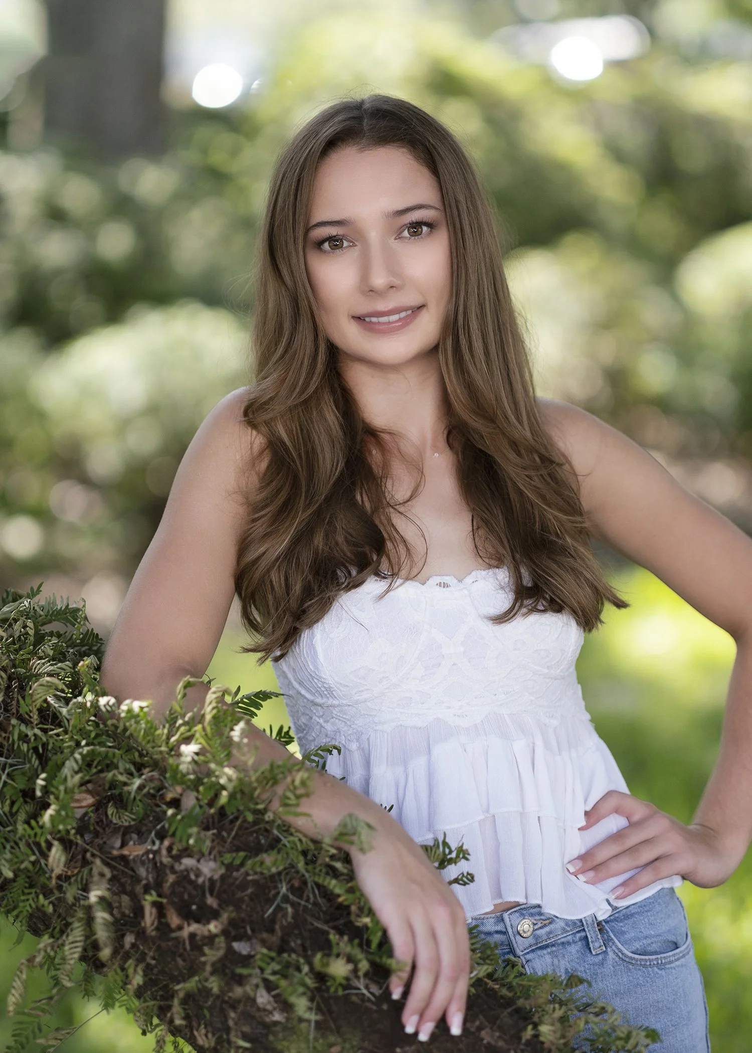 A young woman with long brown hair, wearing a white sleeveless top and blue jeans, stands outdoors in a garden with greenery. She is outdoors with a blurred background of trees and sunlight.
