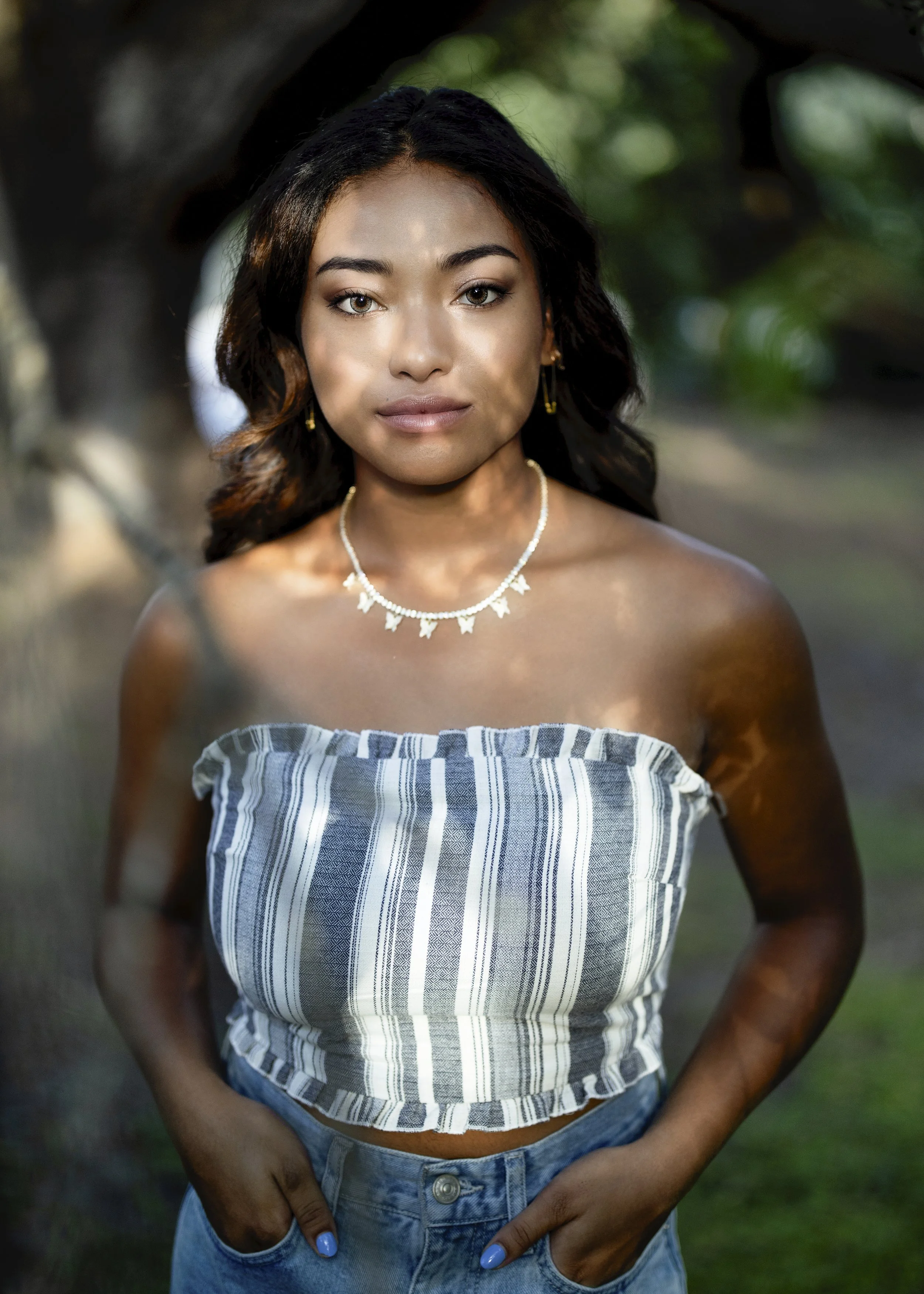 Close-up of a young woman with dark wavy hair, wearing a striped off-shoulder top, a necklace, and earrings, standing outdoors with a blurred background of trees and greenery.