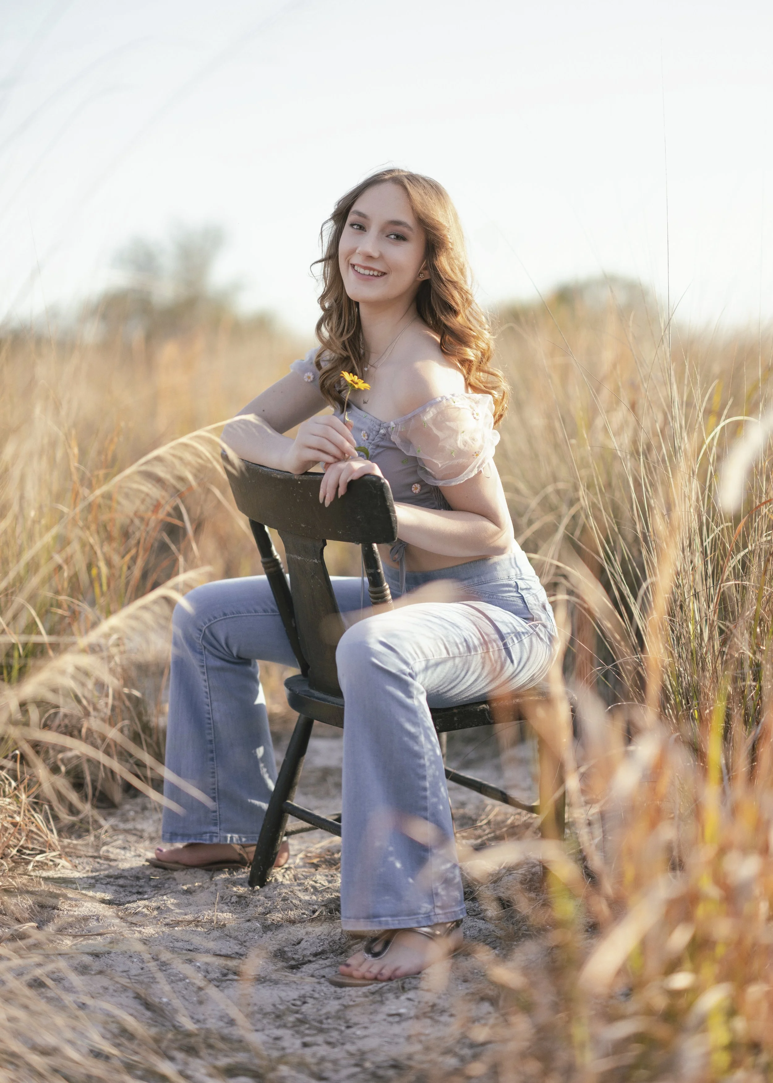 A young woman with long, curly hair smiling and sitting on a black chair in a grassy field holding a yellow flower.