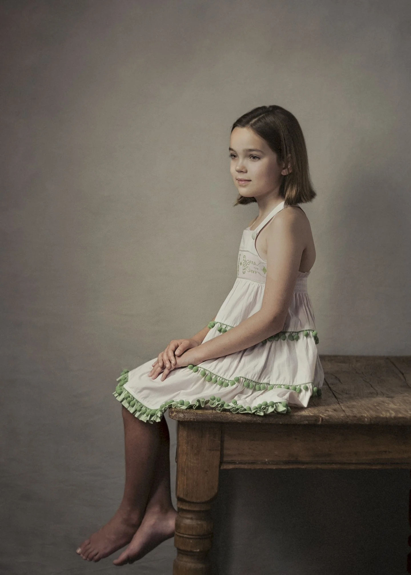 A young girl with shoulder-length brown hair sitting on a wooden stool, wearing a white dress with green pom-pom trim, against a plain gray background.