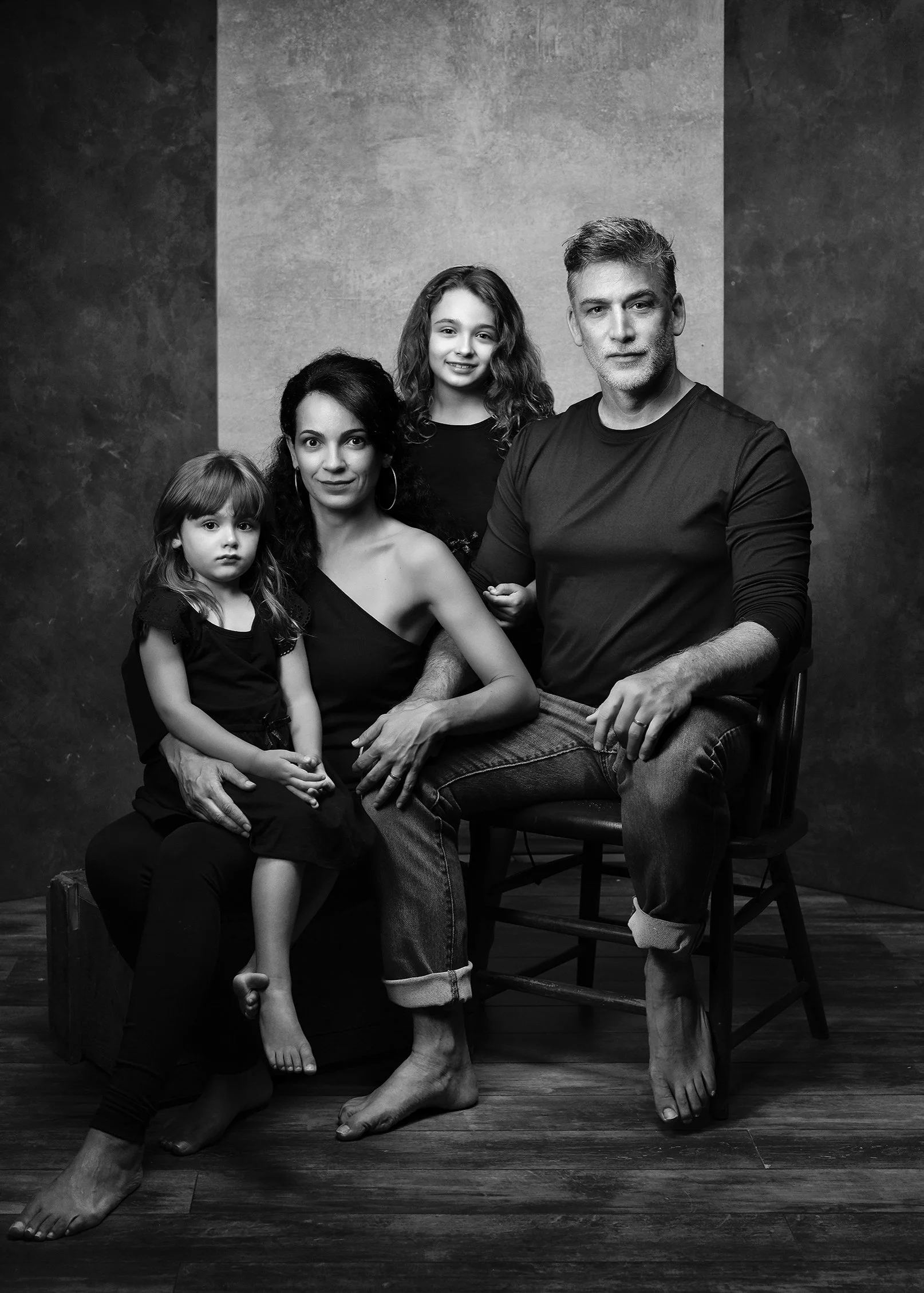 Black and white photograph of a family of four, sitting and standing in a studio. The mother, with curly hair, is sitting on a chair, the father, with short hair, is sitting on a chair with his arm resting on his knee, and the children, a young girl 