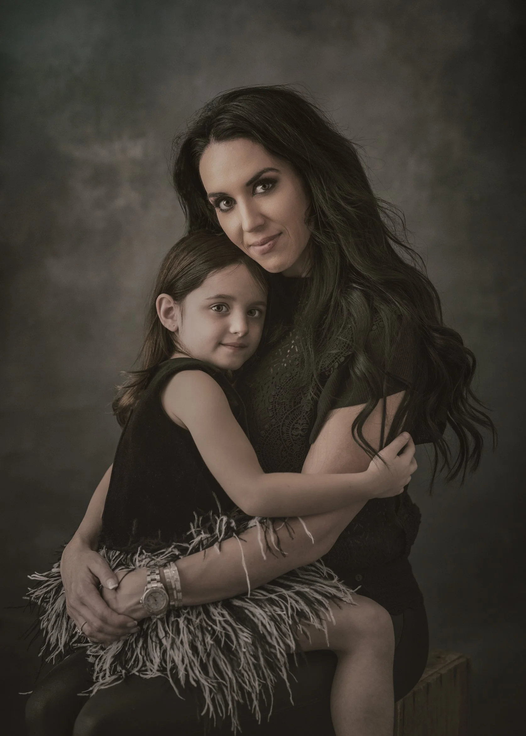 A woman with long dark hair and a young girl with brown hair, both dressed in black, embracing each other in a studio portrait against a dark background.