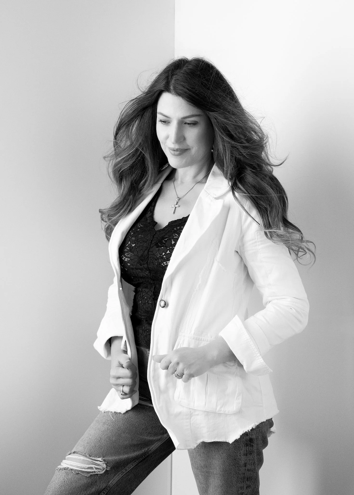Black and white photo of a woman with long, wavy hair wearing a white jacket, black lace top, distressed jeans, and a cross necklace, standing with her hand on her hip.