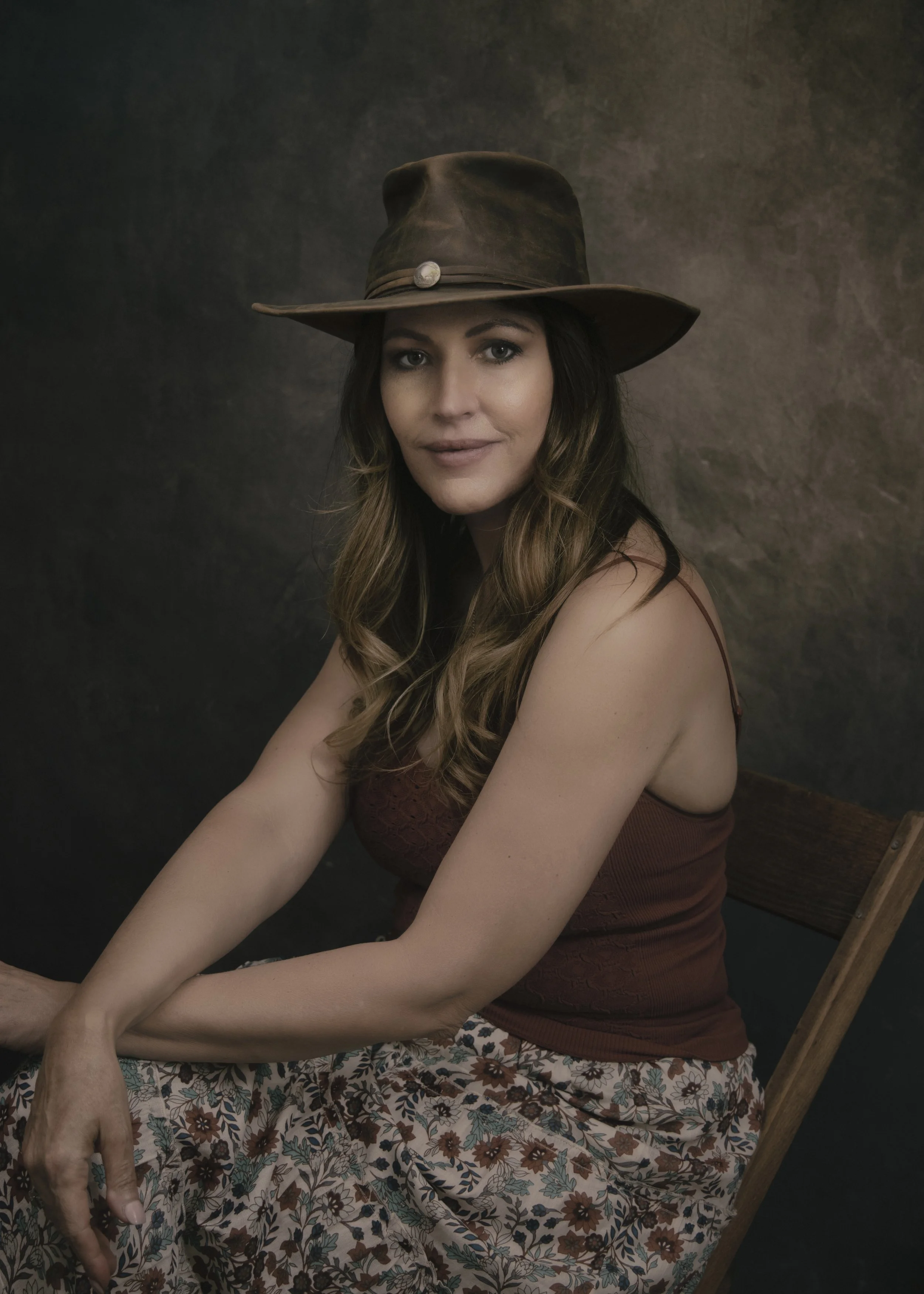 A woman with long wavy hair wearing a brown wide-brimmed hat with a button, a maroon sleeveless top, and a floral skirt, sitting on a wooden chair against a dark textured background.