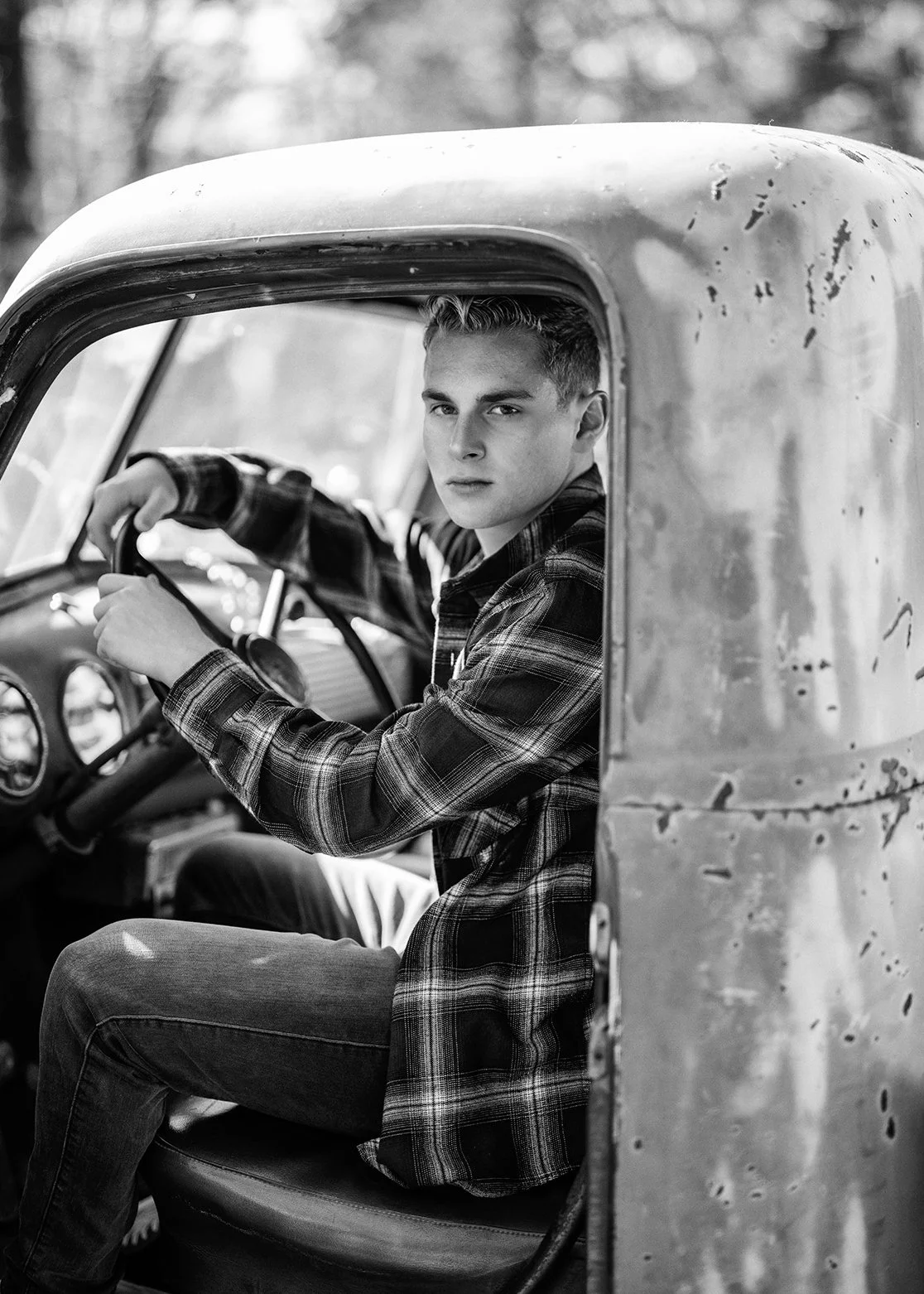 A young man with short hair and wearing a plaid shirt, sitting in the driver’s seat of an old, weathered truck, looking toward the camera.