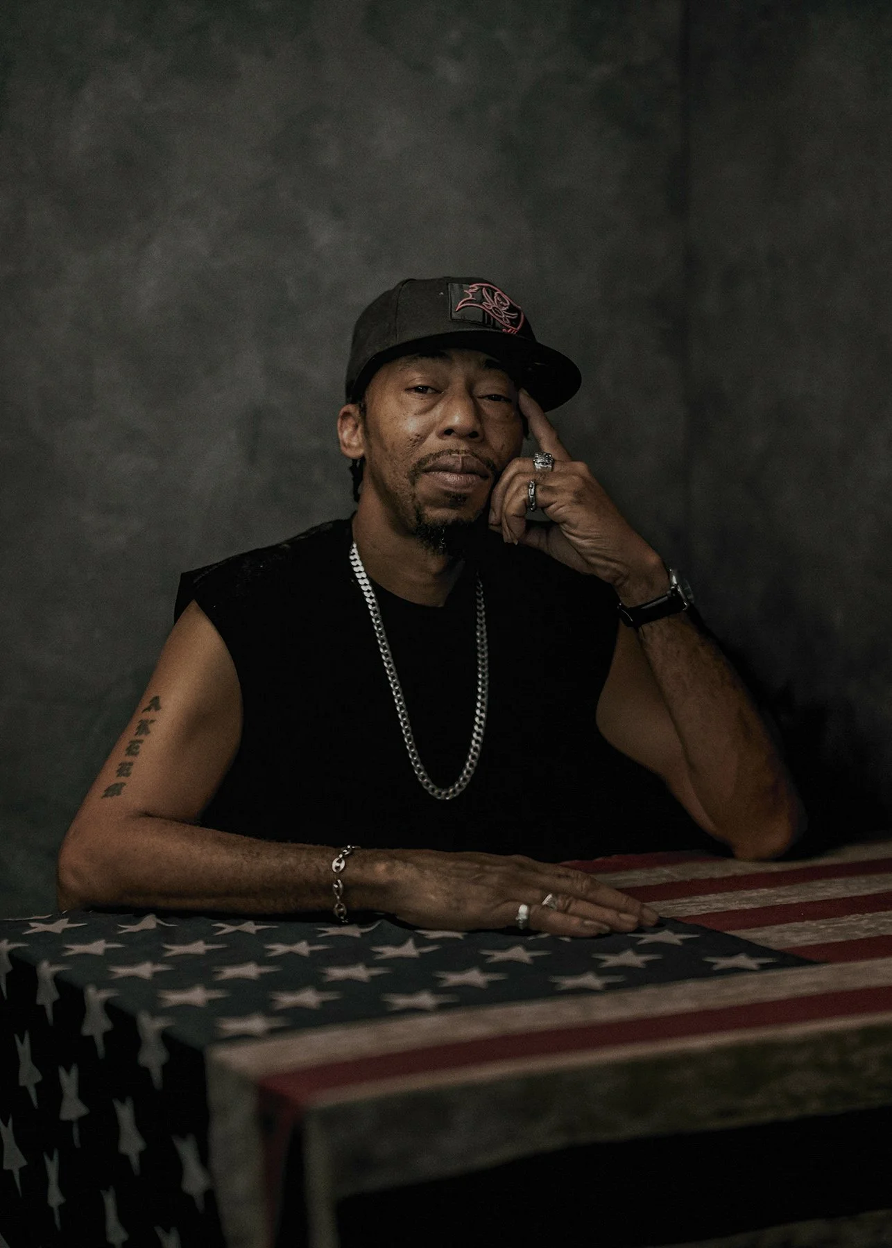 An older man sitting at a table draped with an American flag tablecloth, posing with his hand to his temple, wearing a black sleeveless shirt, a cap, and jewelry, against a dark background.