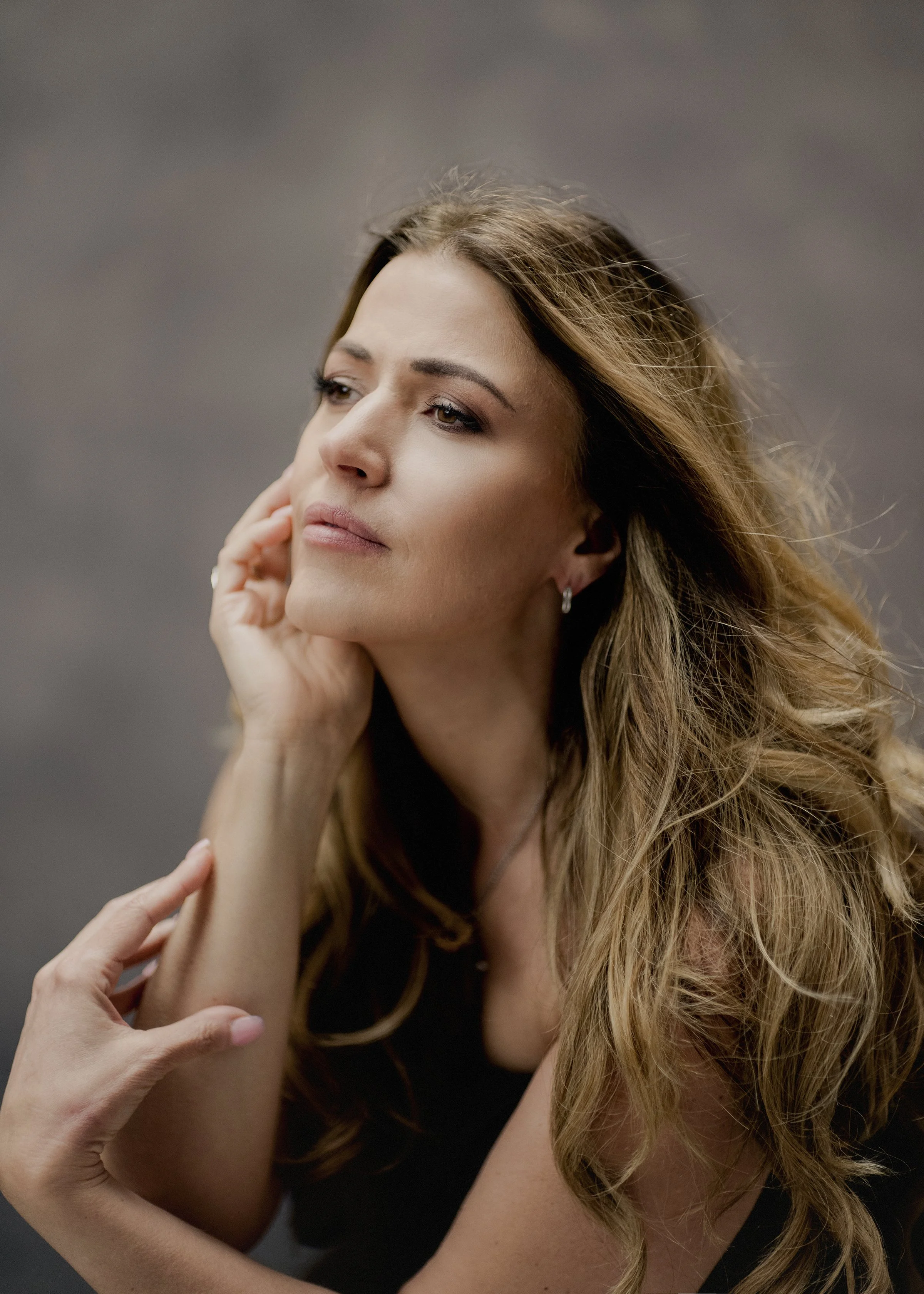 A woman with long, wavy hair resting her chin on her hand, being touched on the arm by another person.