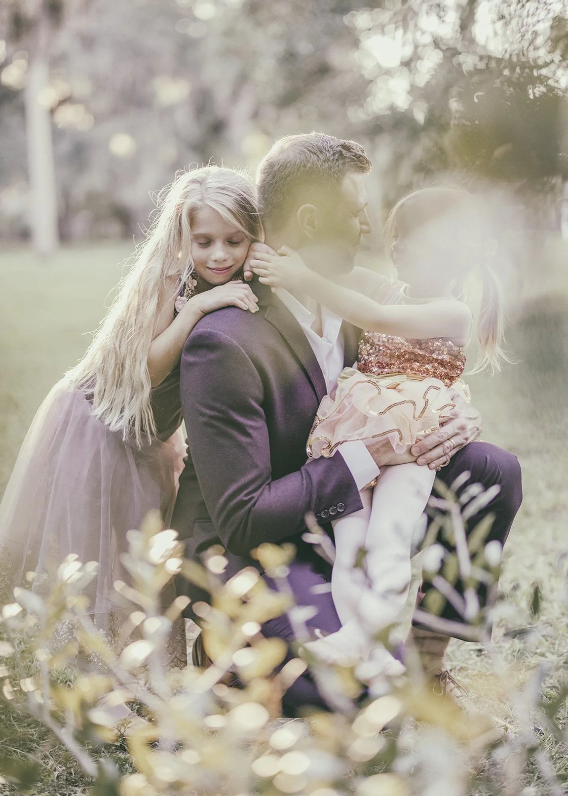 A man in a suit sitting outdoors with two young girls, one hugging him from behind and the other sitting on his lap, all smiling and enjoying a moment in the park during daylight.