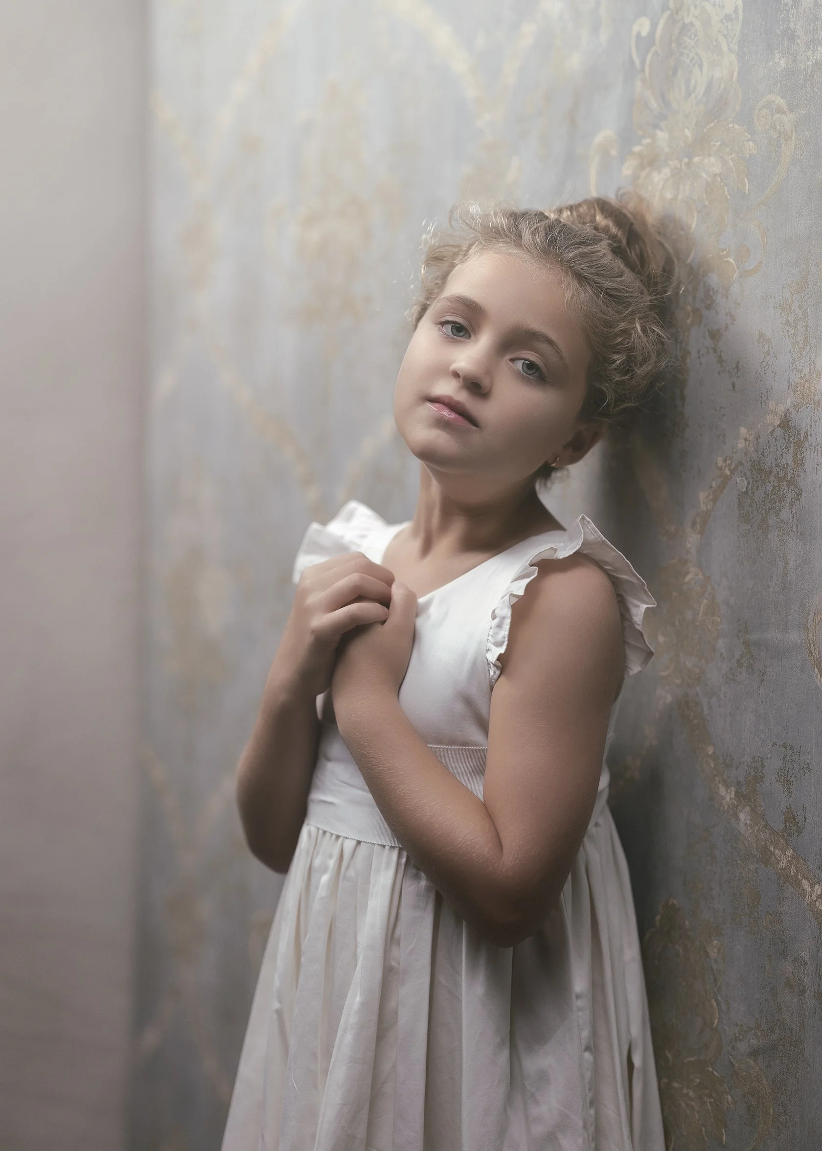 A young girl with curly blonde hair and blue eyes, wearing a white dress with ruffled sleeves, standing against a textured wall with a floral pattern, looking directly at the camera with a serene expression.
