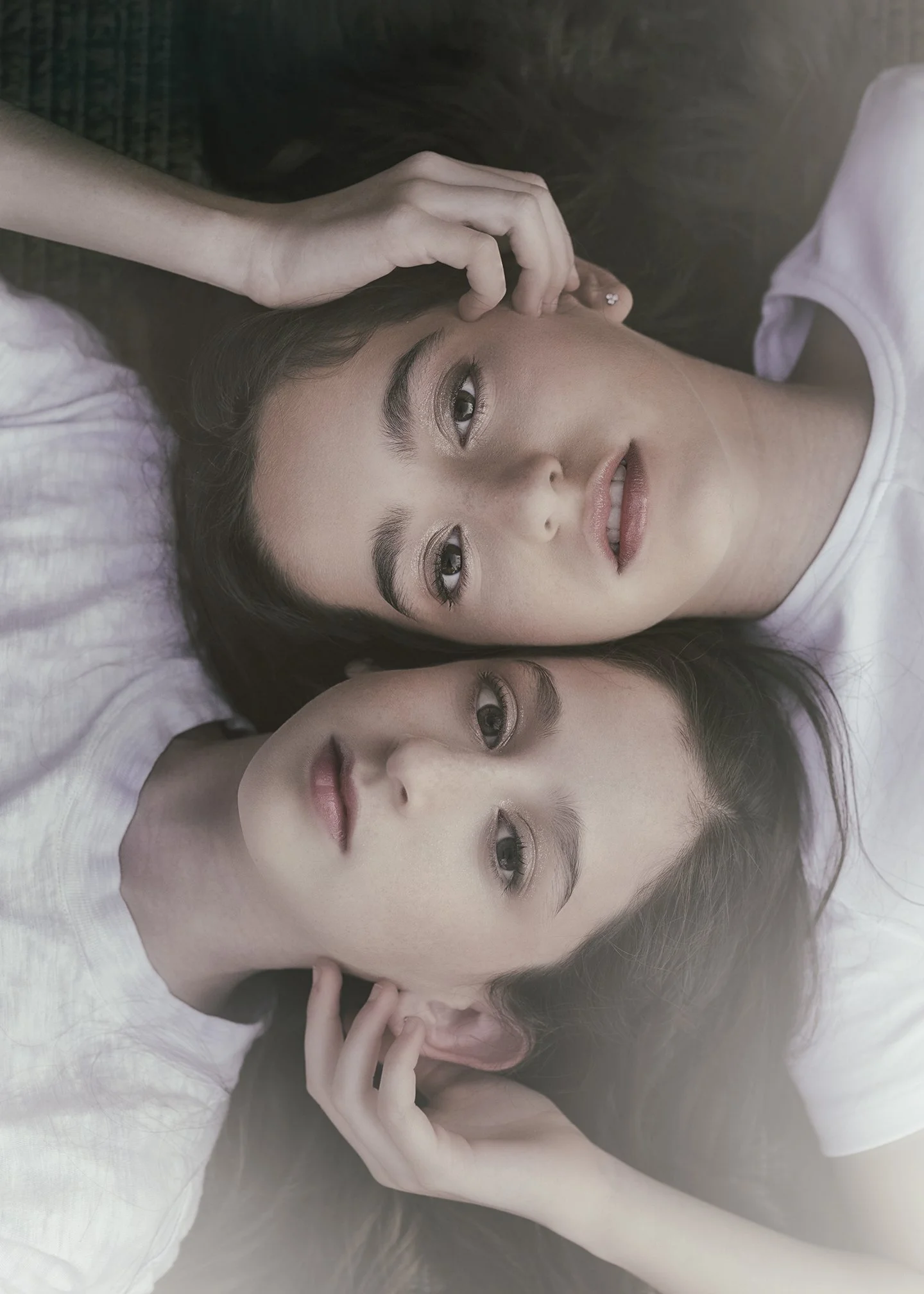 Two young women lying down with heads close together, one with long dark hair and the other with long light hair, both looking at the camera with relaxed expressions.