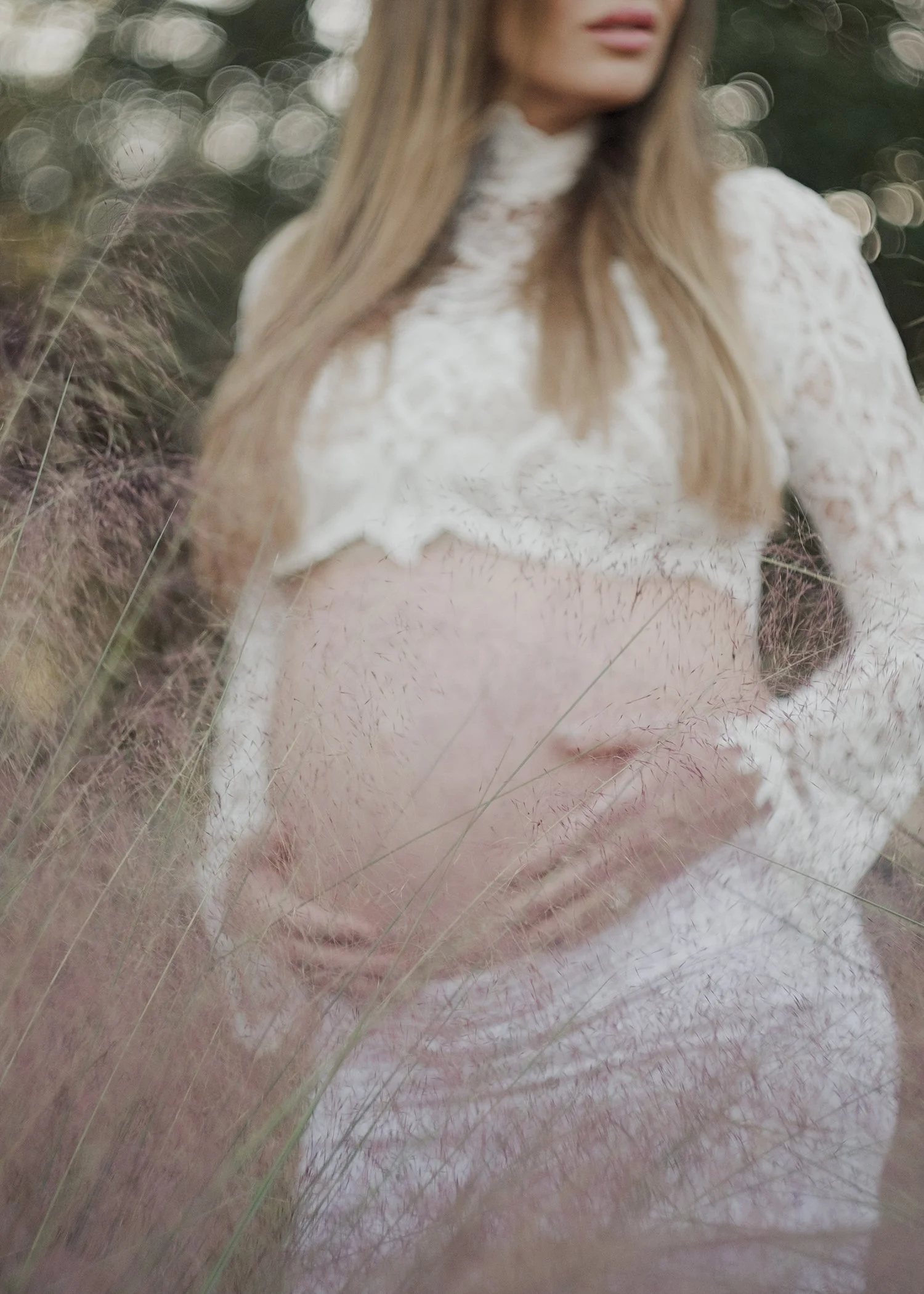 A woman with long blonde hair wearing a white lace top standing outdoors in a field of tall grass, with a blurred background of trees and circular bokeh light effects.