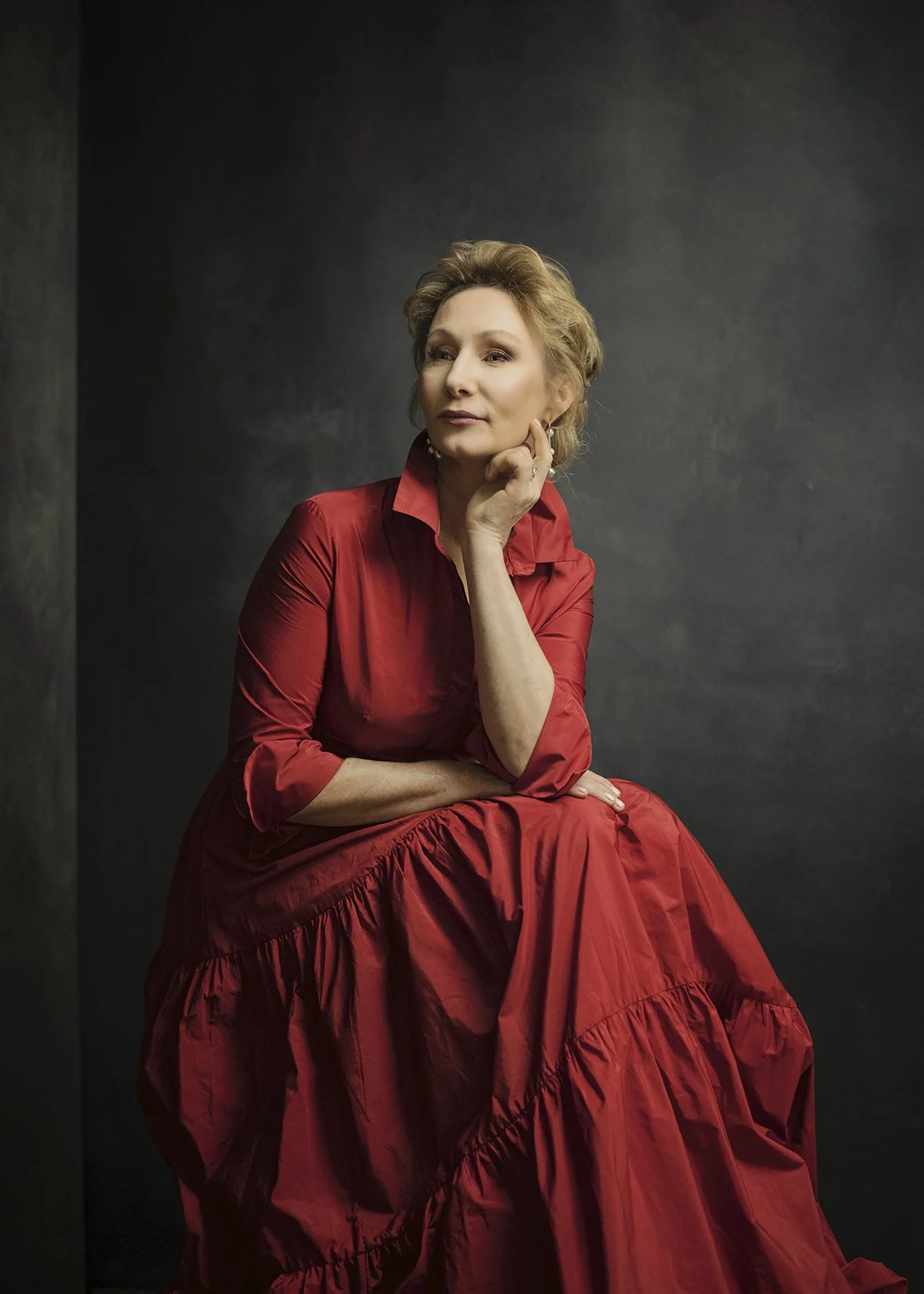 A woman with blonde hair wearing a red dress seated against a dark background, resting her chin on her hand.