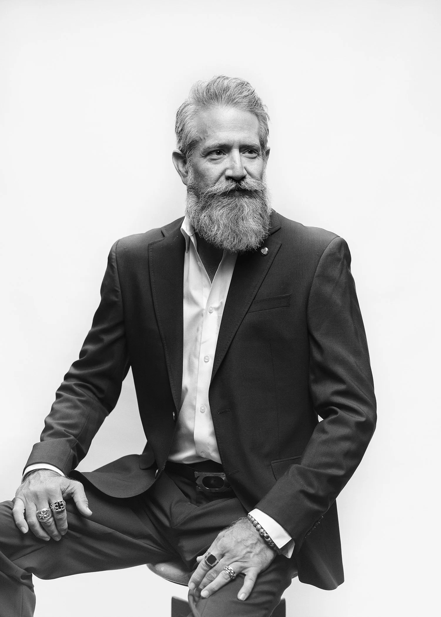 Black and white photo of a man with gray hair and beard, wearing a suit, sitting with crossed legs, and accessorized with rings and bracelets, against a plain background.