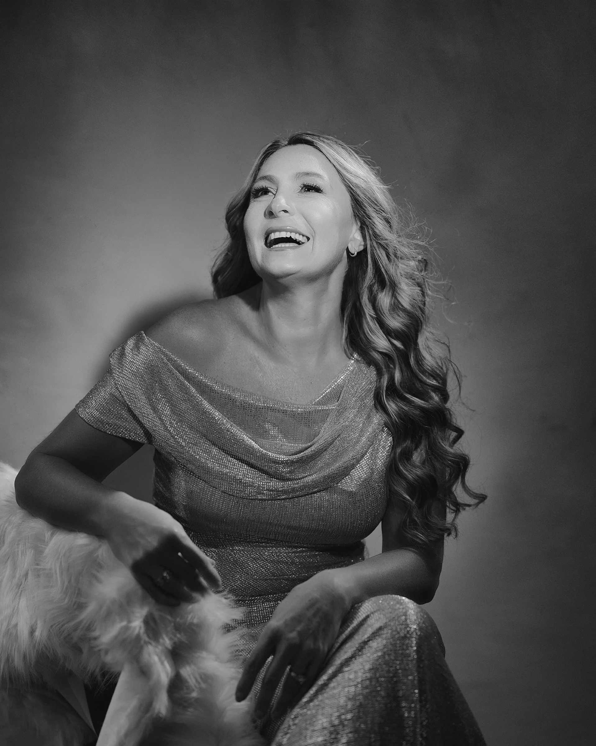 A woman with long curly hair, wearing a shimmering off-the-shoulder dress, laughing and looking slightly upward, sitting beside a furry dog.