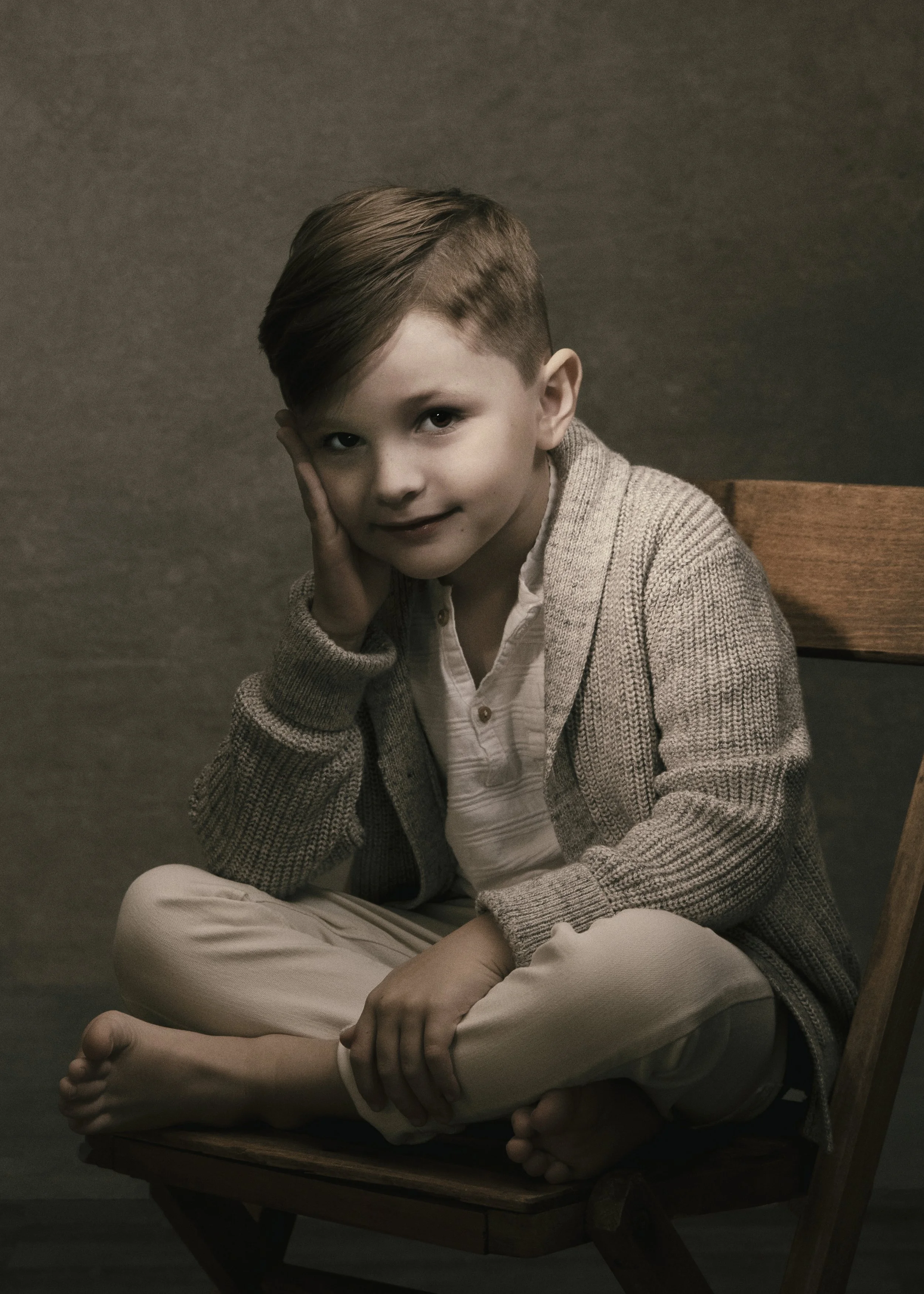 A young boy with brown hair and fair skin sitting on a wooden chair with a gray background. He is wearing a gray sweater over a white shirt and beige pants, sitting cross-legged on the chair. He supports his head with his right hand, resting it on hi