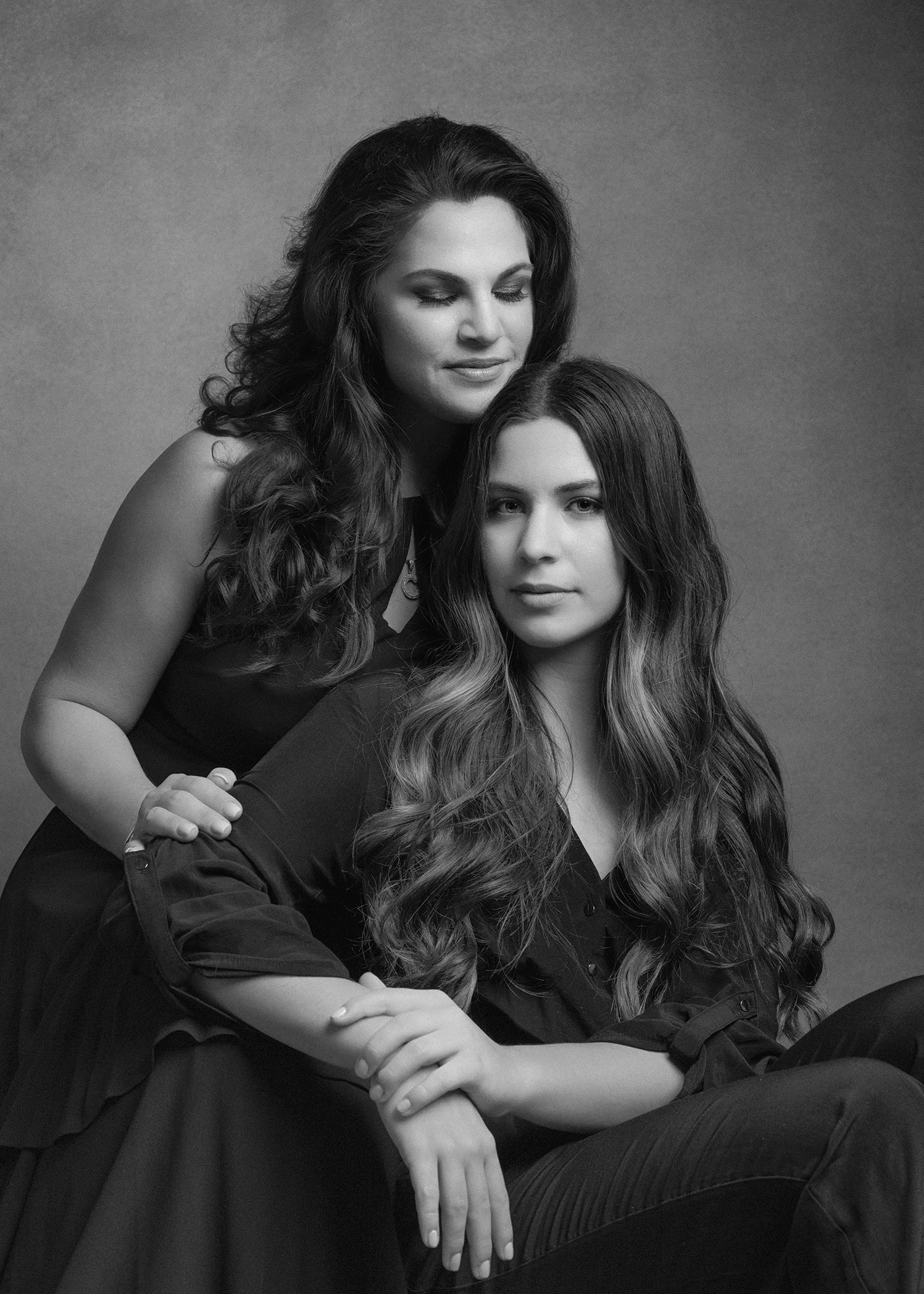 Black and white portrait of two women, one older and one younger, with long, wavy hair. The older woman is standing behind the younger woman, who is seated. Both have calm, serene expressions and their eyes are partially closed.