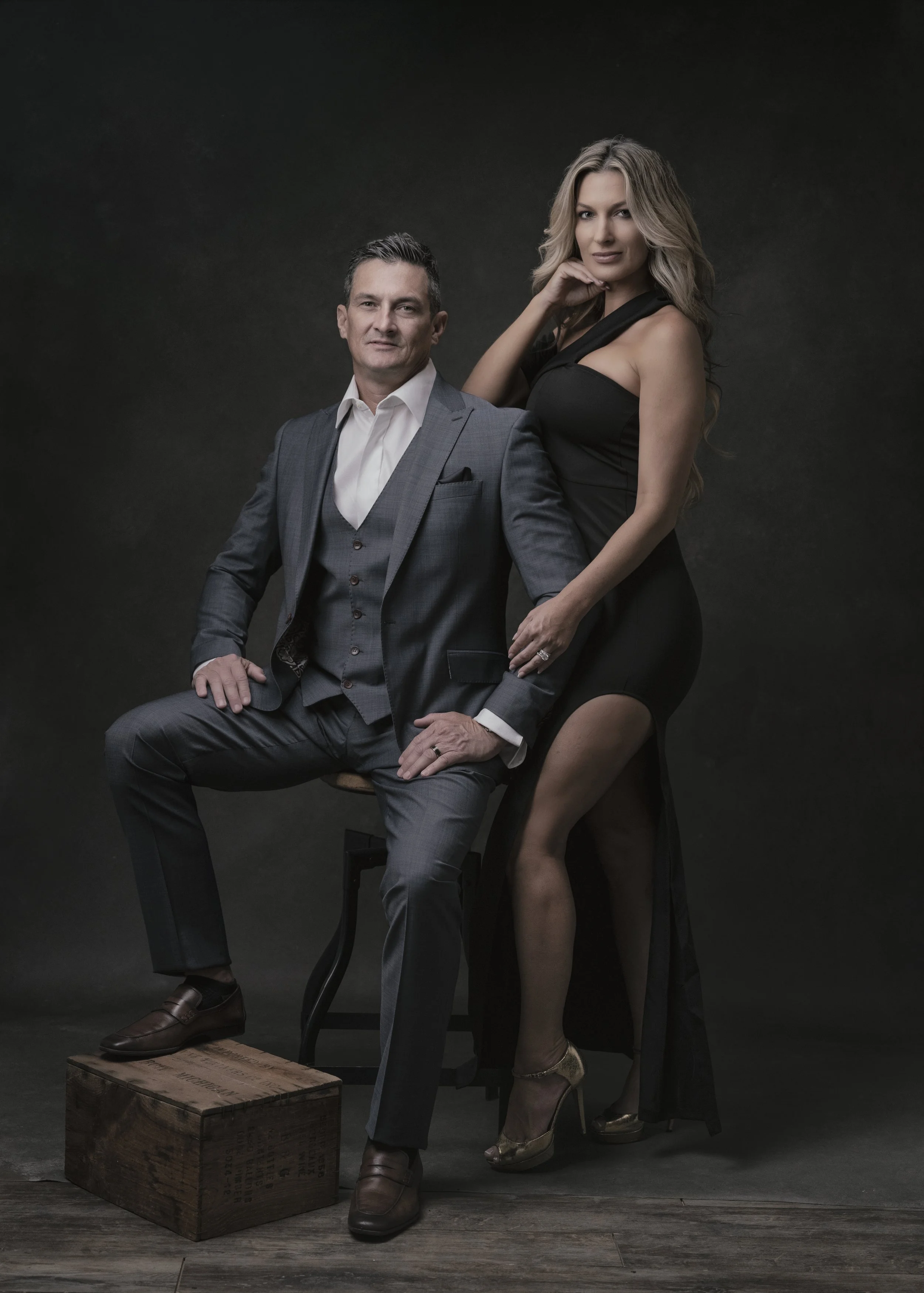 A man and woman pose for a photo against a dark background. The man is seated on a stool, wearing a gray suit, white shirt, and brown shoes. The woman stands behind him, wearing a black dress and gold high heels, with her right hand resting gently on