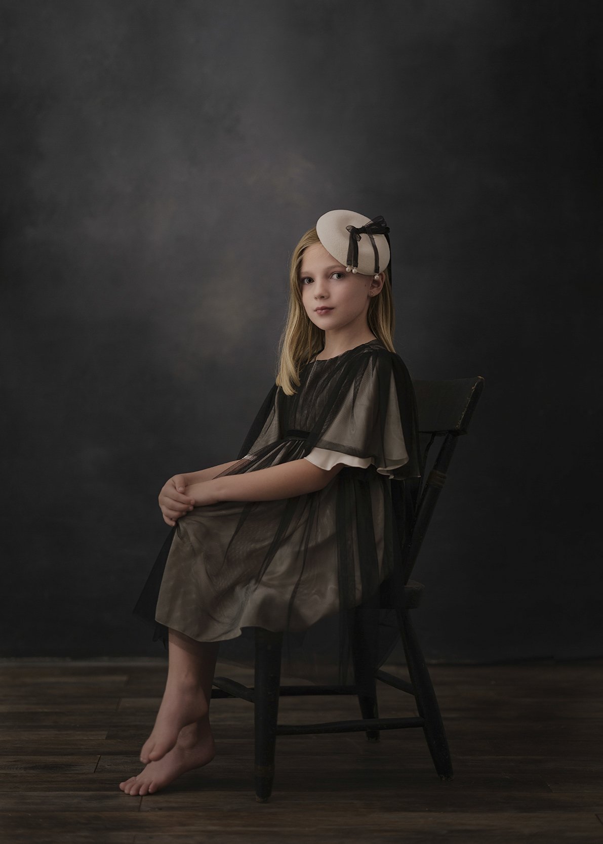 A young girl with blonde hair sitting on a black wooden chair against a dark, textured background. She is wearing a vintage-style dress with sheer black overlay and a beige hat with black ribbon adornment. Her feet are bare, and she is looking slight