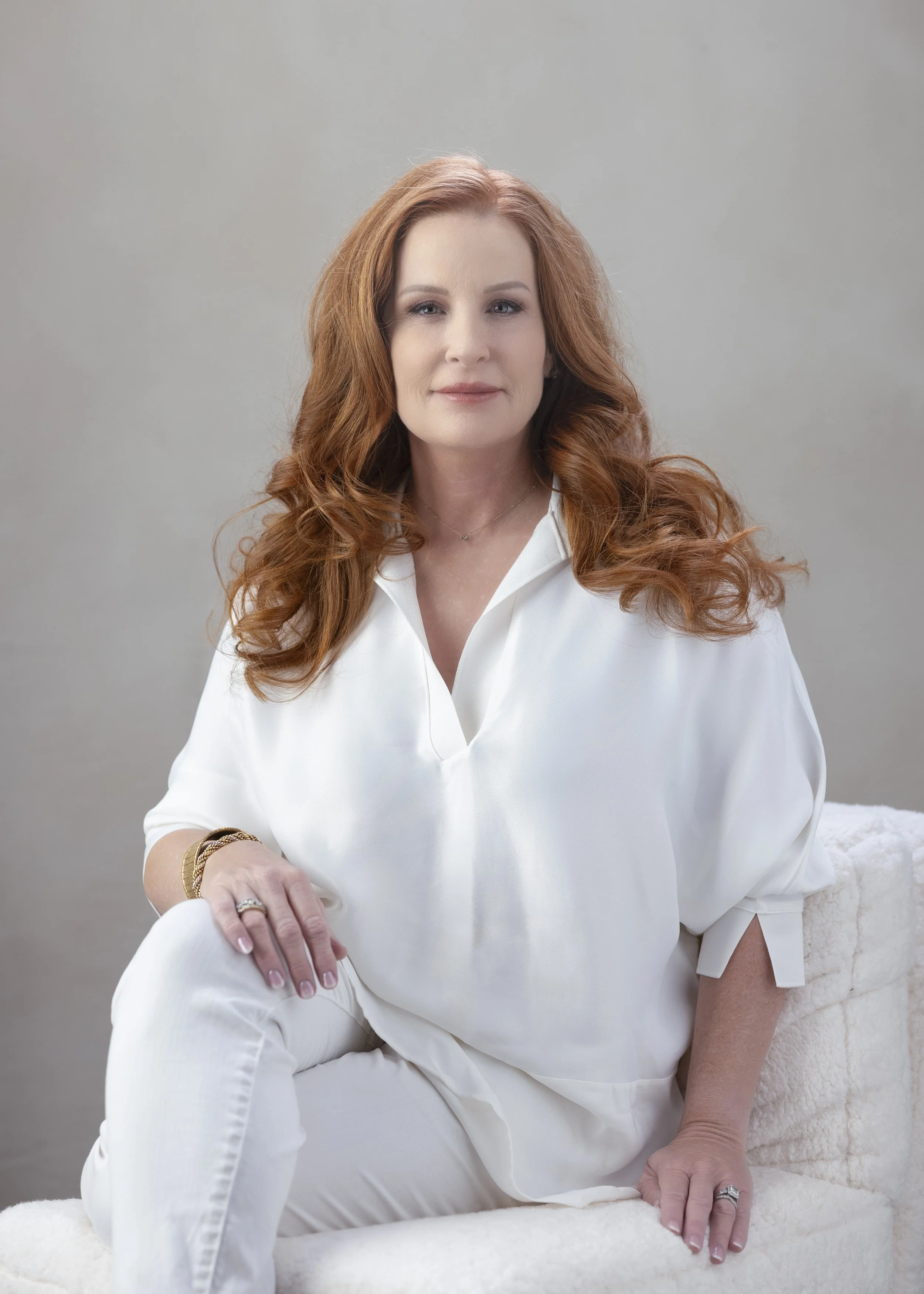 A woman with red wavy hair, wearing a white blouse, sitting on a plush white chair, with a neutral gray background.