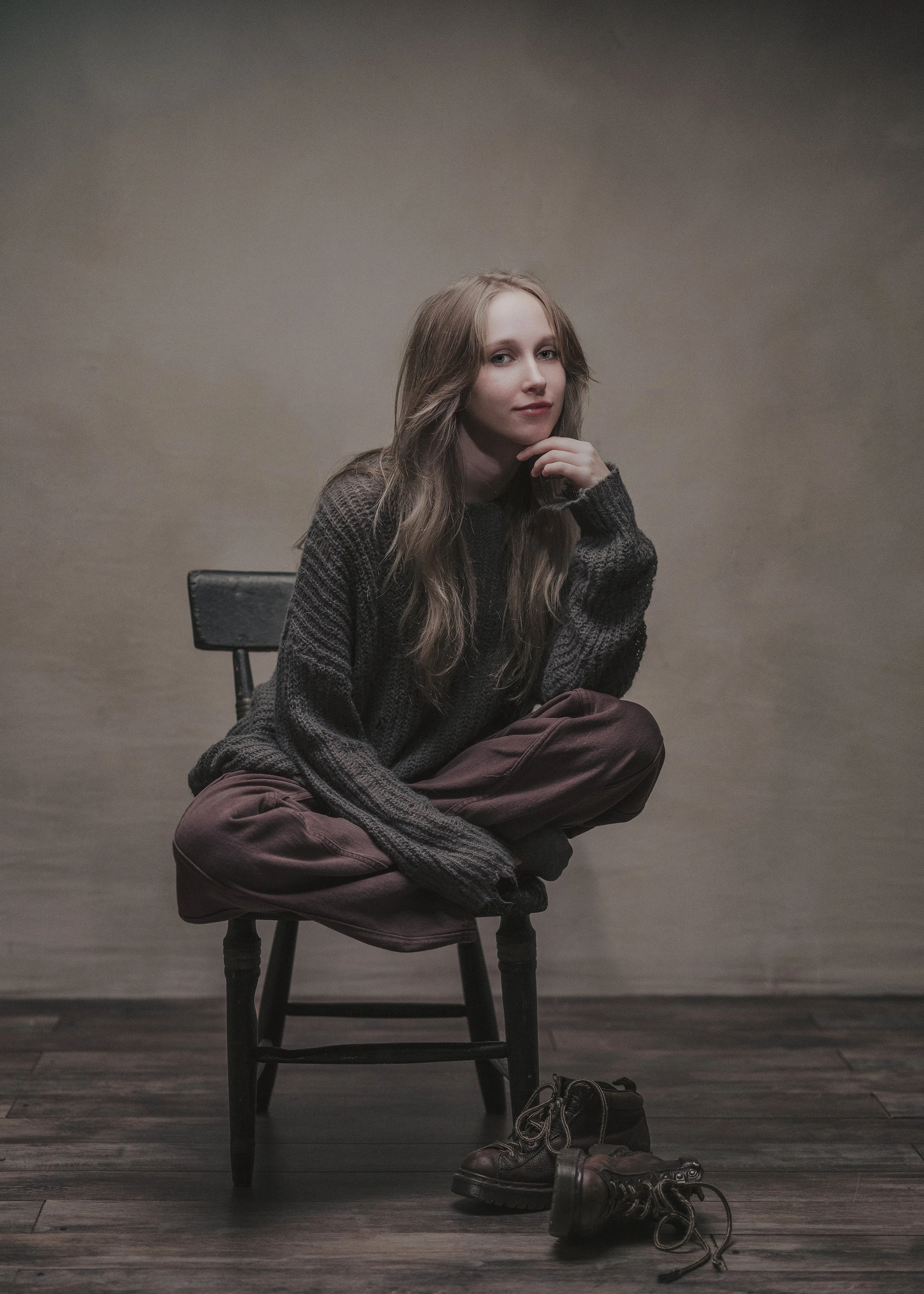 A woman with long light brown hair sitting on a wooden chair against a plain neutral background. She is wearing a dark sweater and brown pants, with black boots placed on the floor in front of her. Her left hand rests near her chin and she looks dire