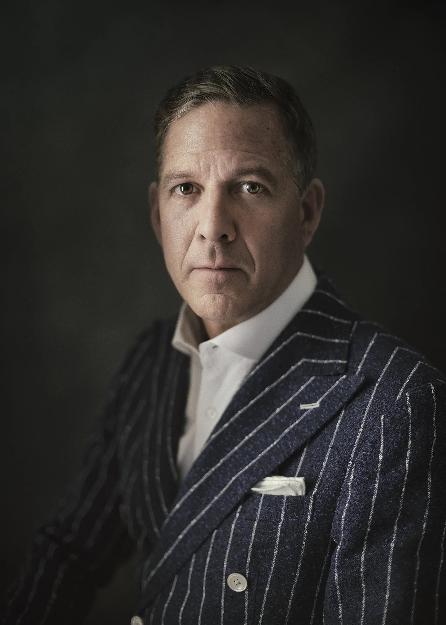 A man with short, dark hair, wearing a white dress shirt and a dark navy blazer with white pinstripes, against a dark background.
