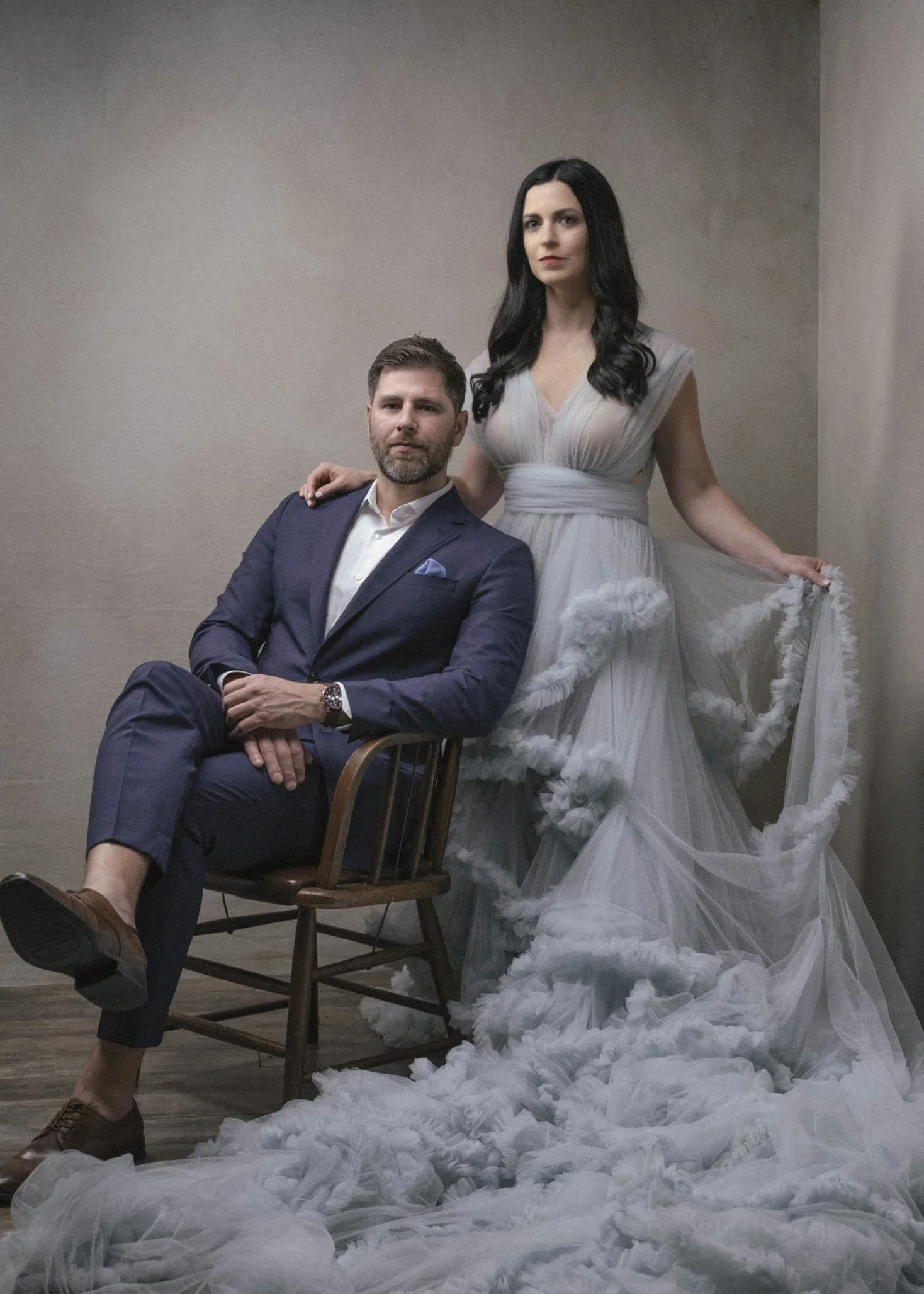 A man in a navy suit and white shirt sitting on a wooden chair with his legs crossed, and a woman in a flowing light-colored dress standing behind him with her hand on his shoulder, in front of a plain wall, surrounded by white fluffy material on the