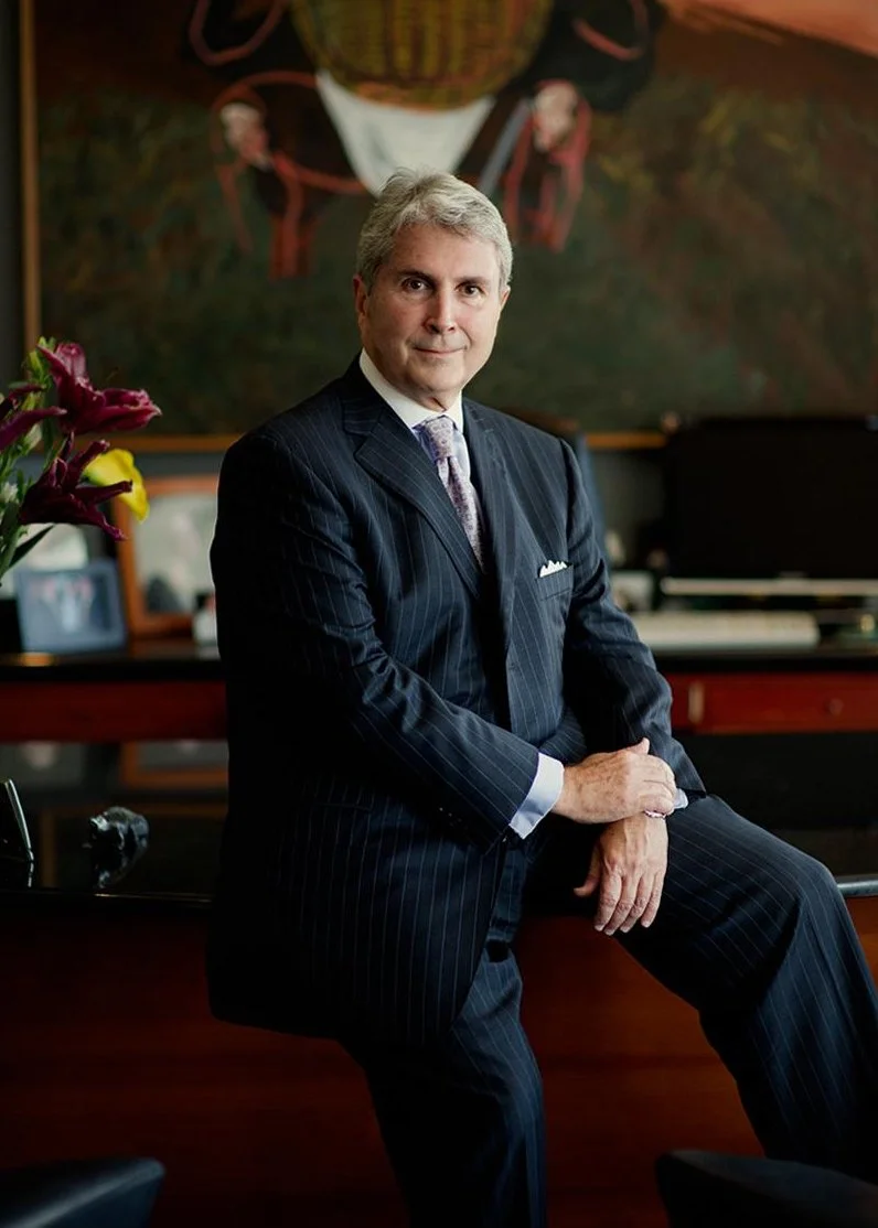 A man in a dark pinstripe suit sitting on a desk in an office or music room, with a colorful painting in the background and flowers on a table nearby.