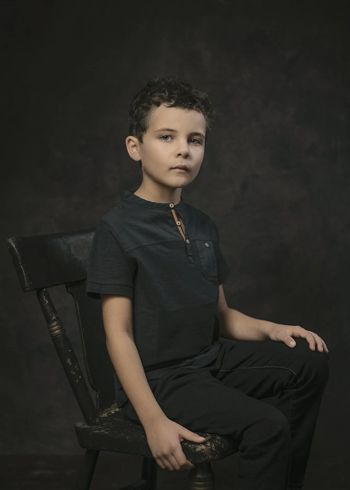 A young boy with curly hair sitting on a black wooden chair against a dark background, wearing a black shirt and pants.