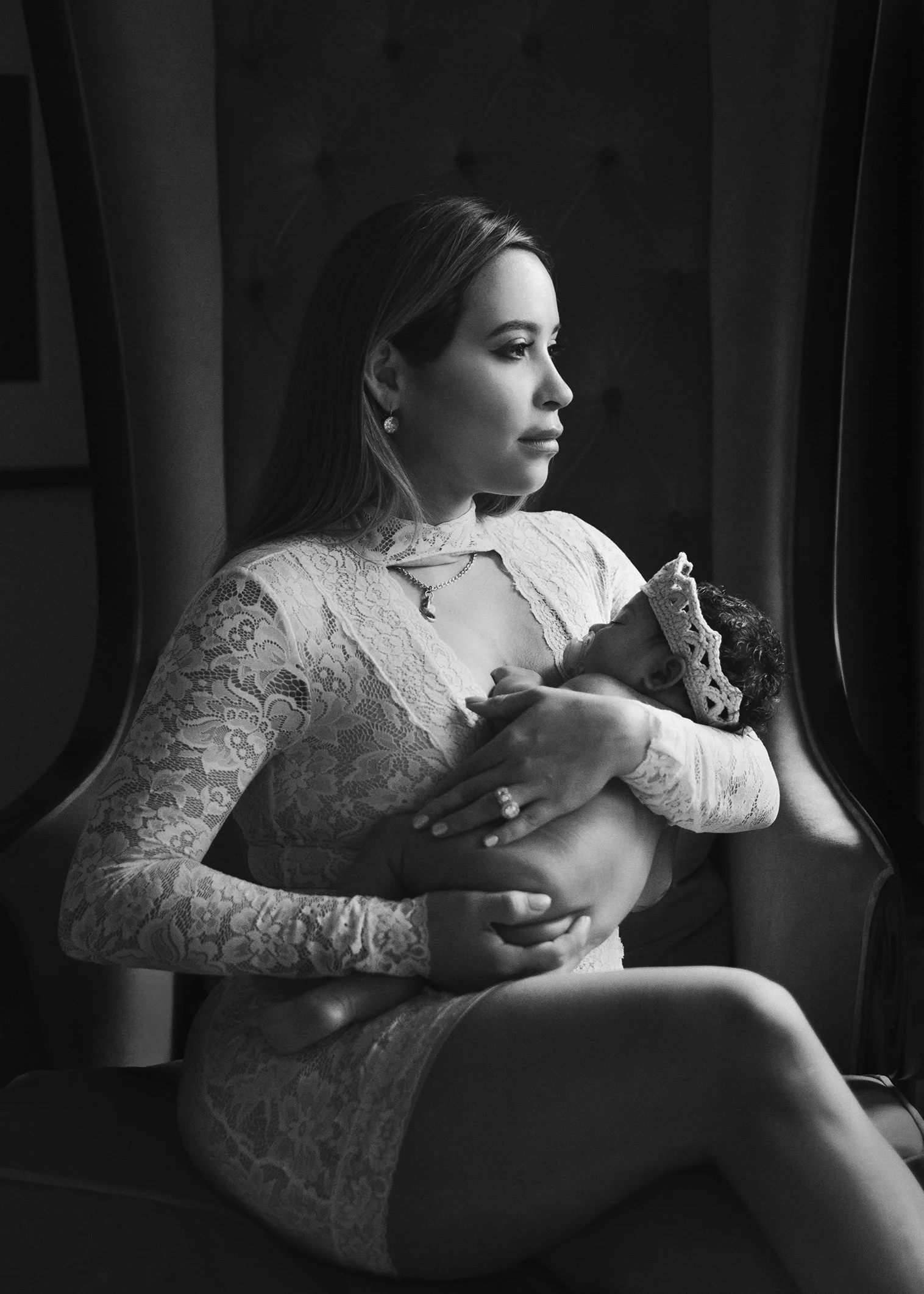 A woman with long hair, wearing a lace dress and earrings, sitting on a train or bus, holding a baby wrapped in a blanket. The woman is looking through a window.