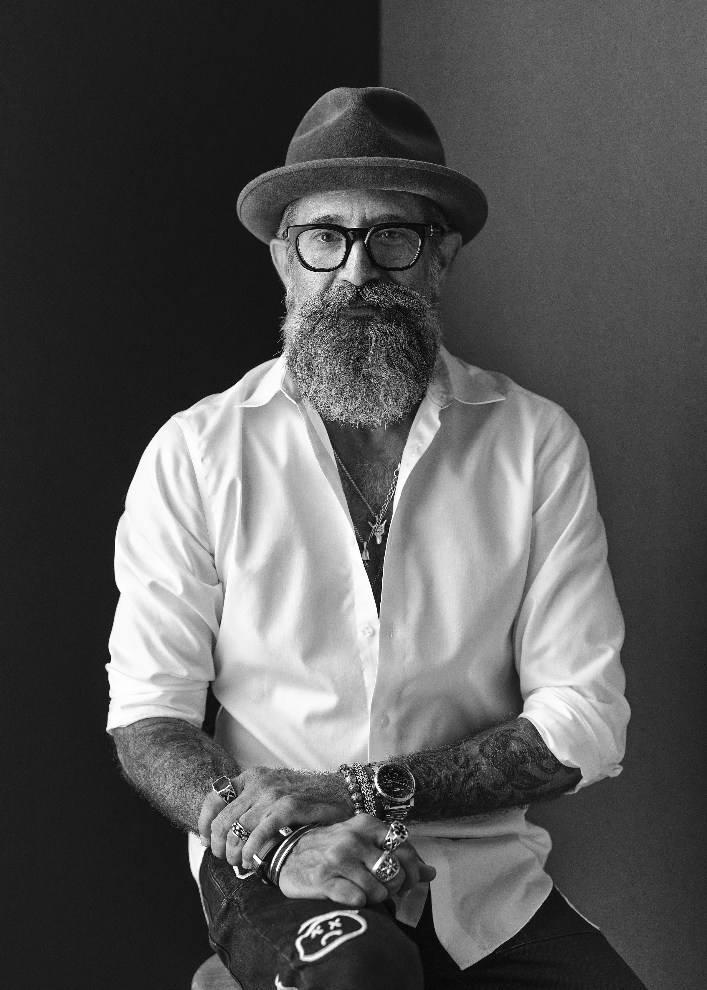 Black and white portrait of a man with a beard and mustache wearing glasses, a fedora hat, a white shirt, and multiple rings and bracelets, sitting with his arms crossed in front of a dark background.