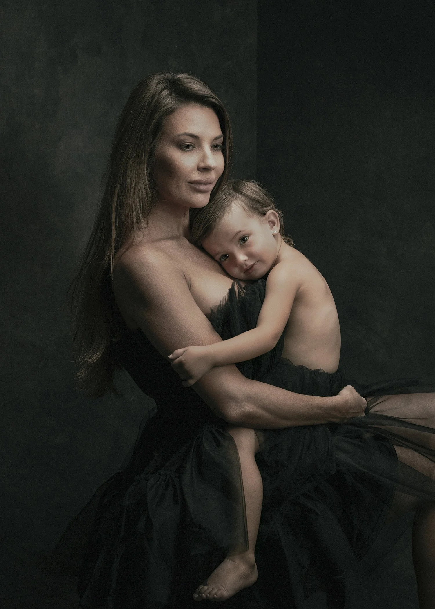 A woman with long brown hair holding a young girl with light brown hair against a black background. The girl is dressed in black and resting her head on the woman's shoulder.