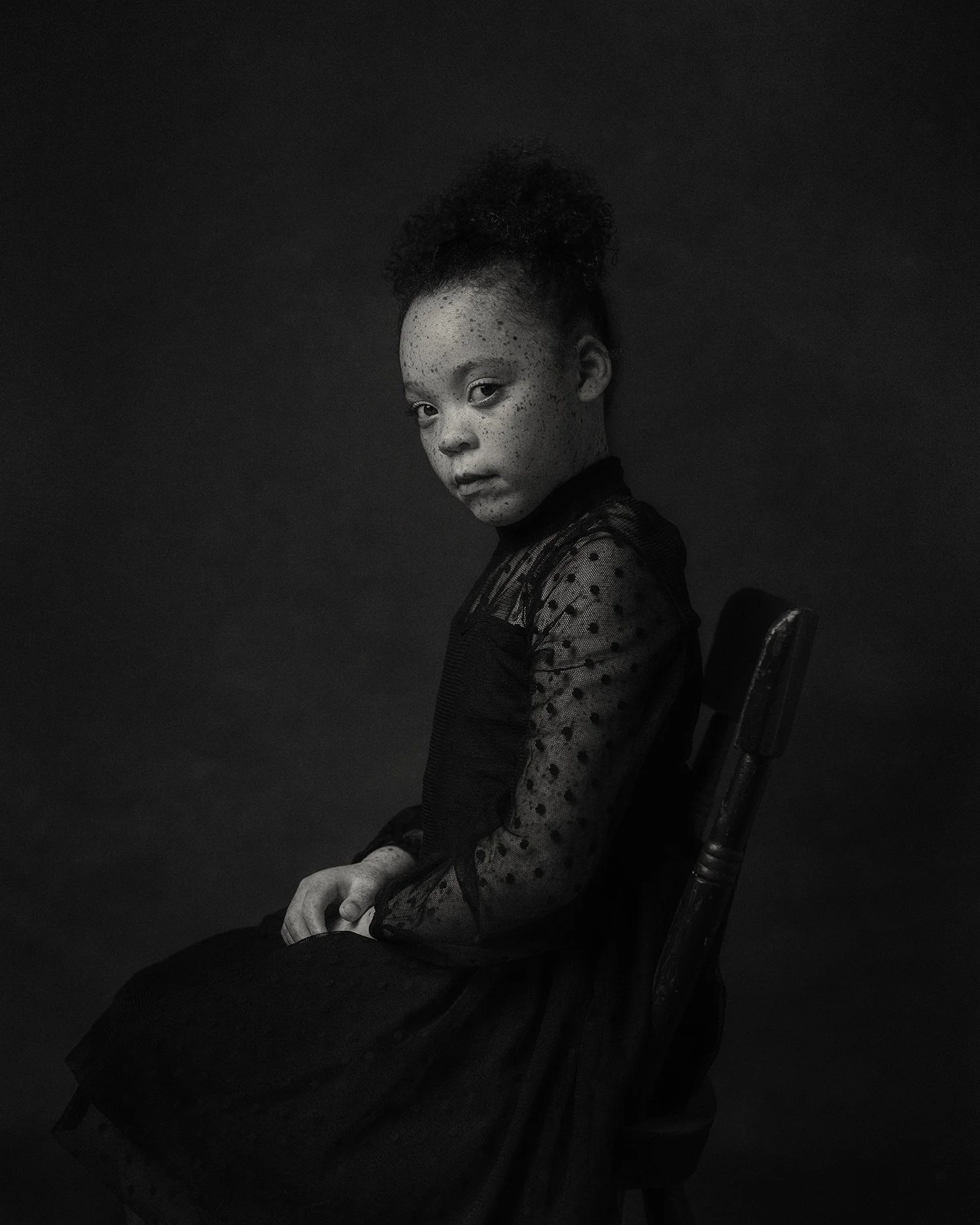 A black and white portrait of a young girl with freckles, curly hair tied up, sitting on a dark wooden chair, wearing a sheer polka dot top.