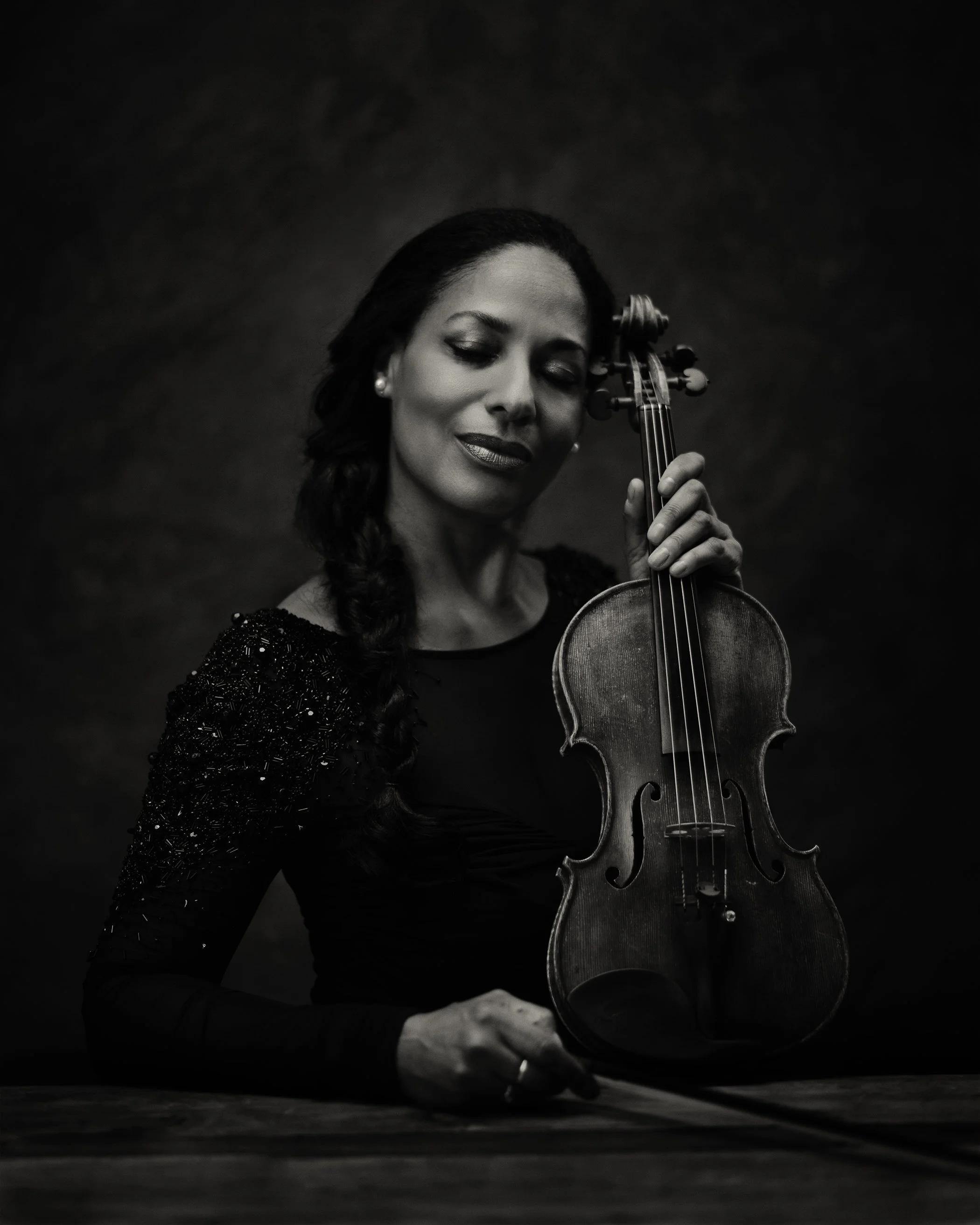 Black and white photo of a woman with closed eyes holding a violin with one hand, her face close to the violin, against a dark background.