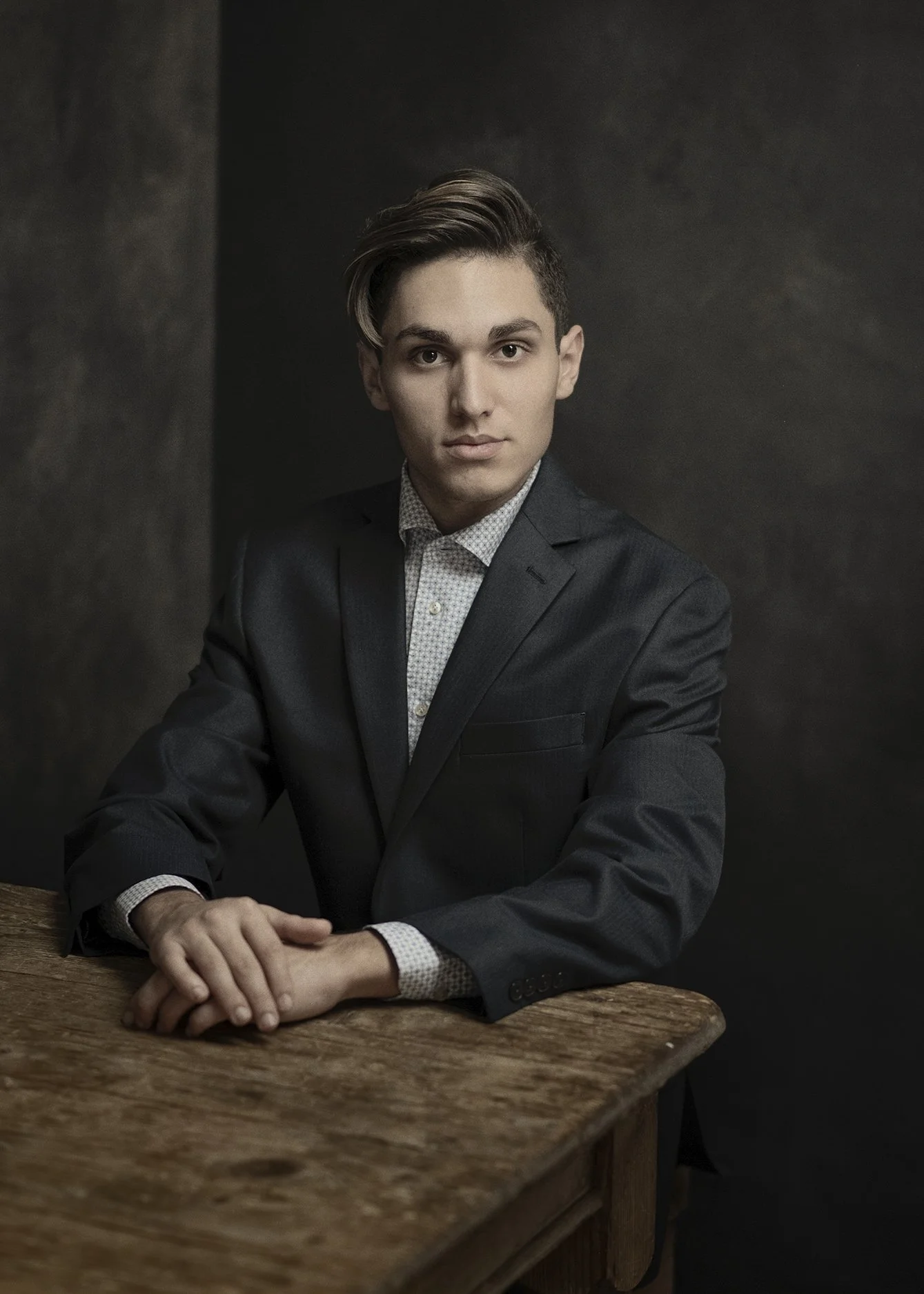 A young man with dark hair styled to the side, wearing a dark suit and white shirt, sitting at a wooden table with his hands clasped, against a dark background.
