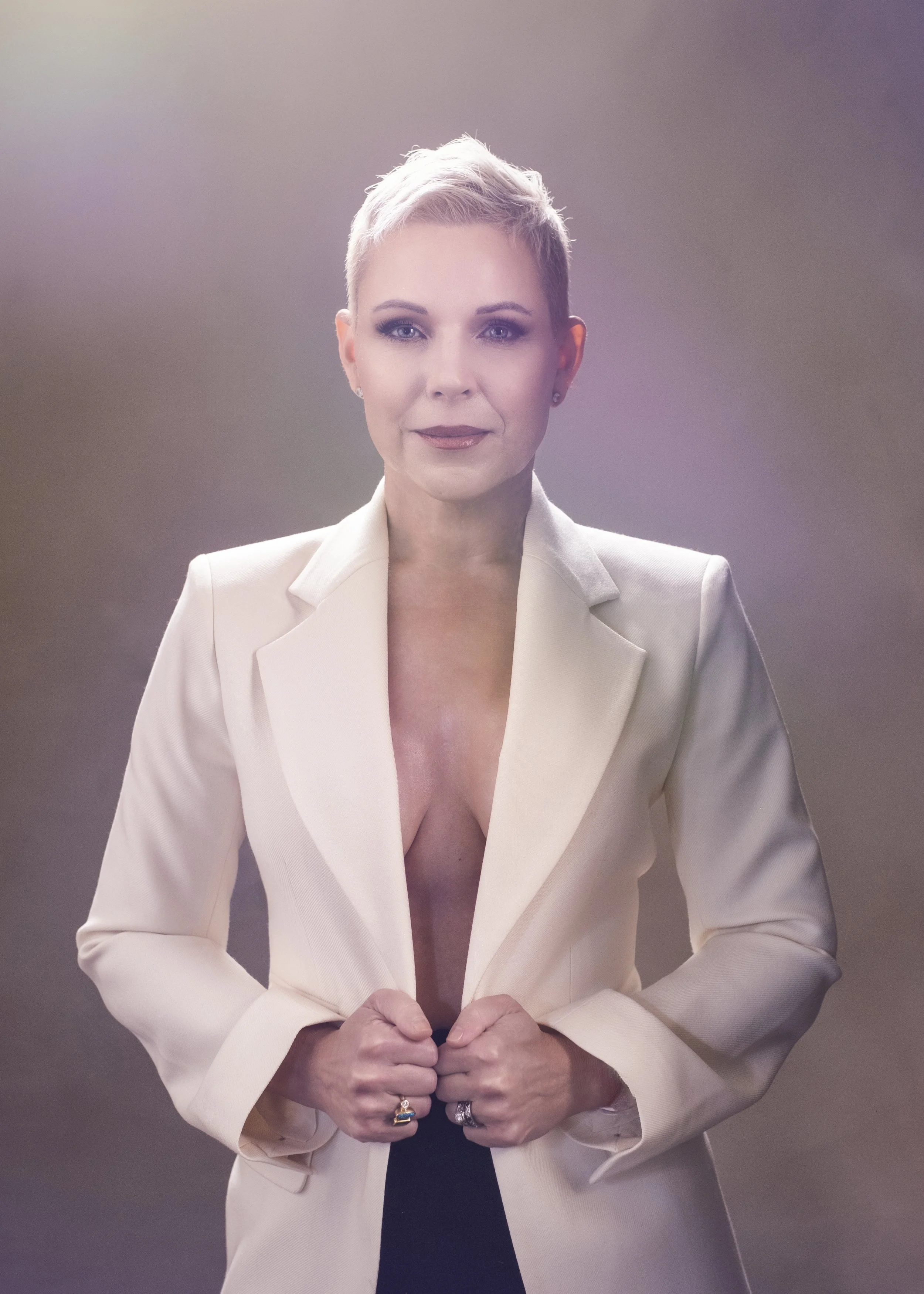 A woman with a short blonde hairstyle wearing a cream-colored blazer and black top, standing with her fists clenched.