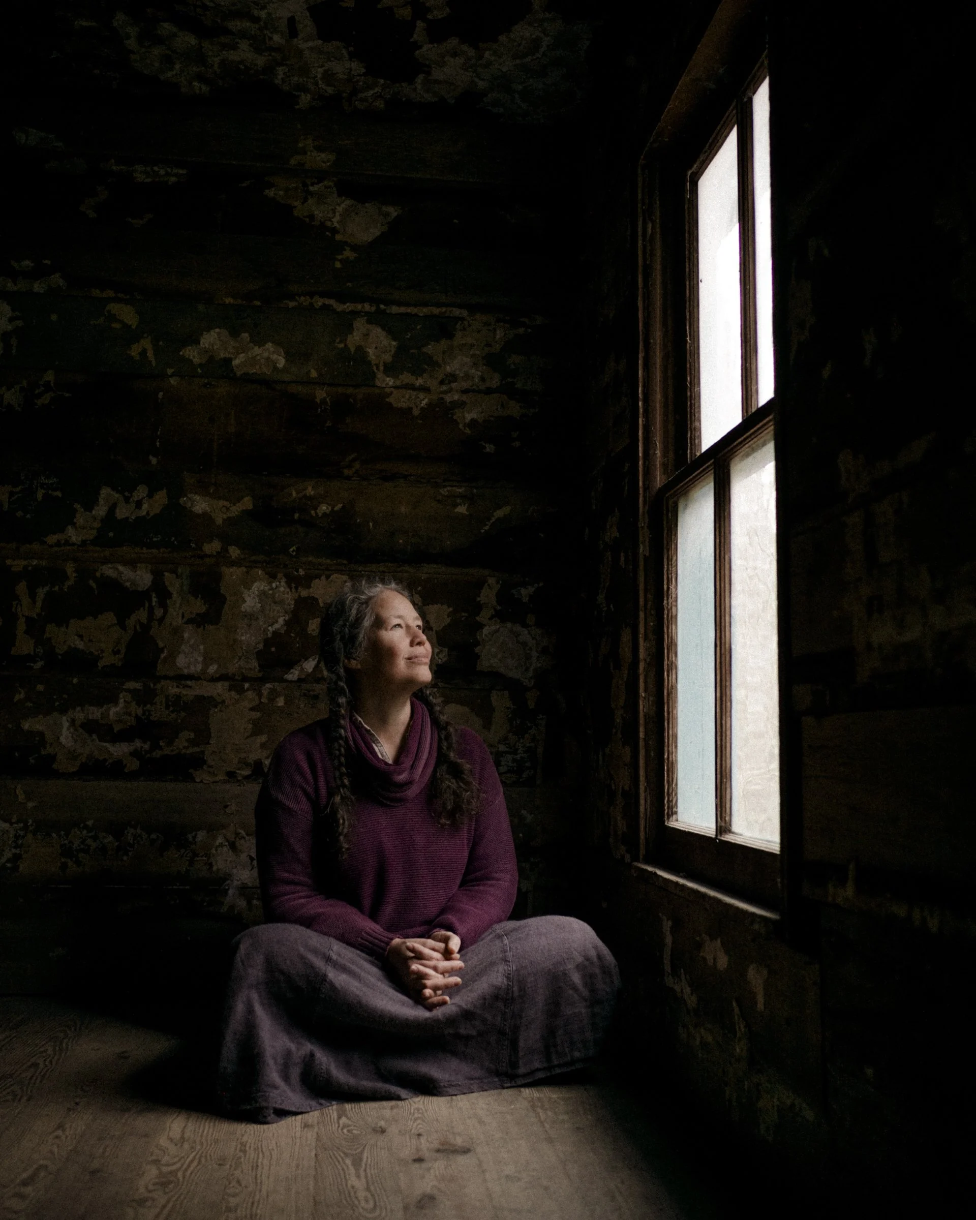 A woman with long braided hair, wearing a dark purple hoodie and gray pants, sitting cross-legged on a wooden floor in a dimly lit room with dark, peeling walls. She is looking thoughtfully toward the window.