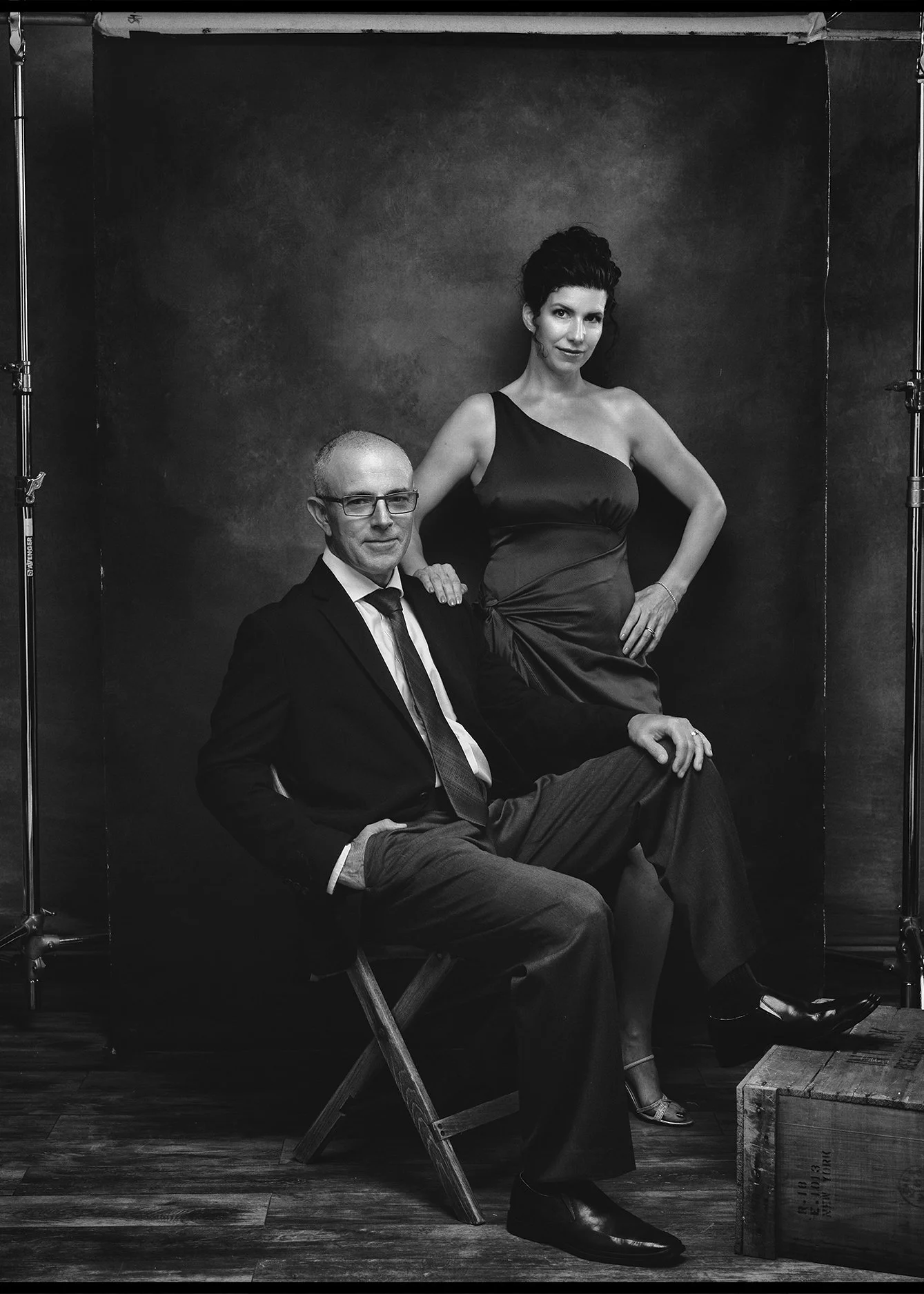 A black and white photograph of a man and woman in formal attire. The man is seated on a director's chair, wearing a suit and glasses, with hand resting on his knee. The woman stands behind him, her hand on his shoulder, wearing a one-shoulder dress 