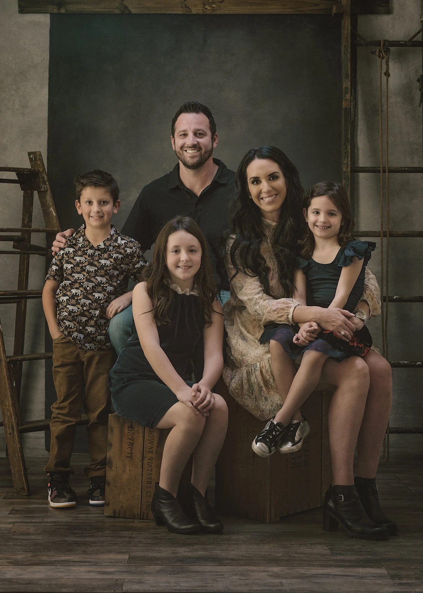 A family photo of six people, including two adults and four children, sitting and standing in front of a dark backdrop with industrial-style ladders and ropes. The family members are smiling and dressed casually.