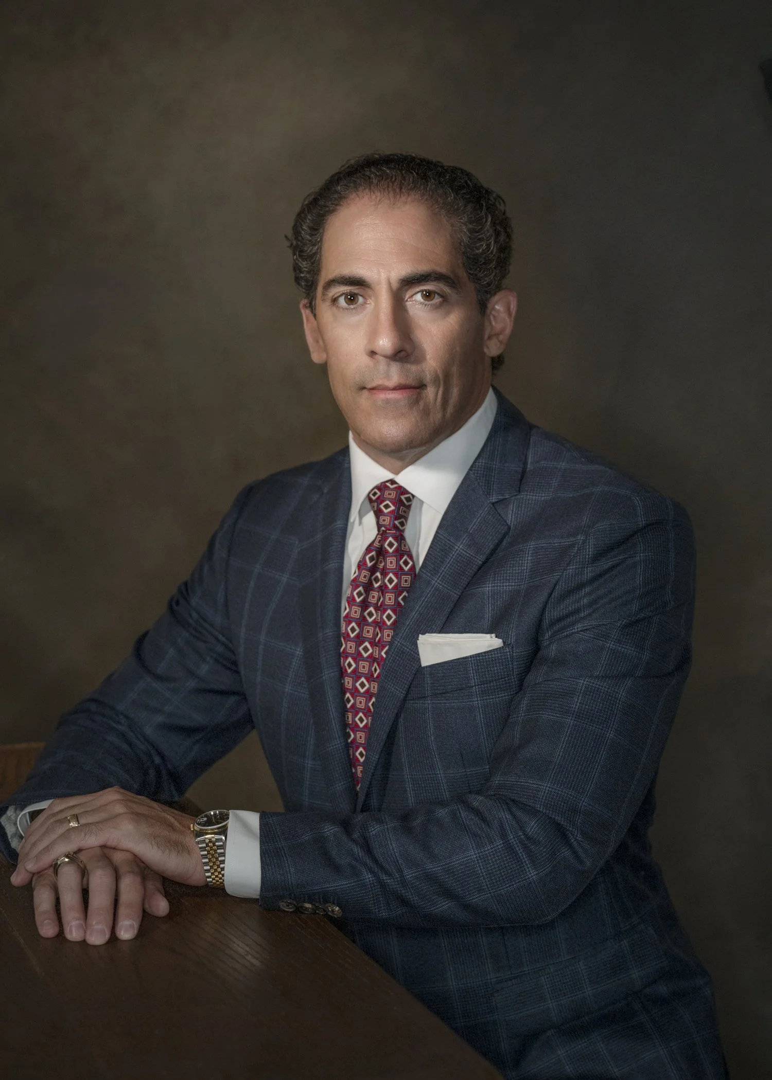 Portrait of a middle-aged man with dark, curly hair, wearing a dark plaid suit, white shirt, and red patterned tie, sitting at a wooden table with hands folded, against a dark brown backdrop.