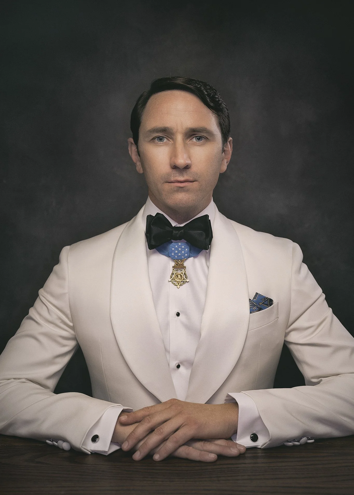 A man in a white tuxedo with a black bow tie sitting at a dark wooden table, with his hands clasped in front of him. He is wearing a medal with a star pendant around his neck, a blue cummerbund, and a blue and gold pocket square. The background is da
