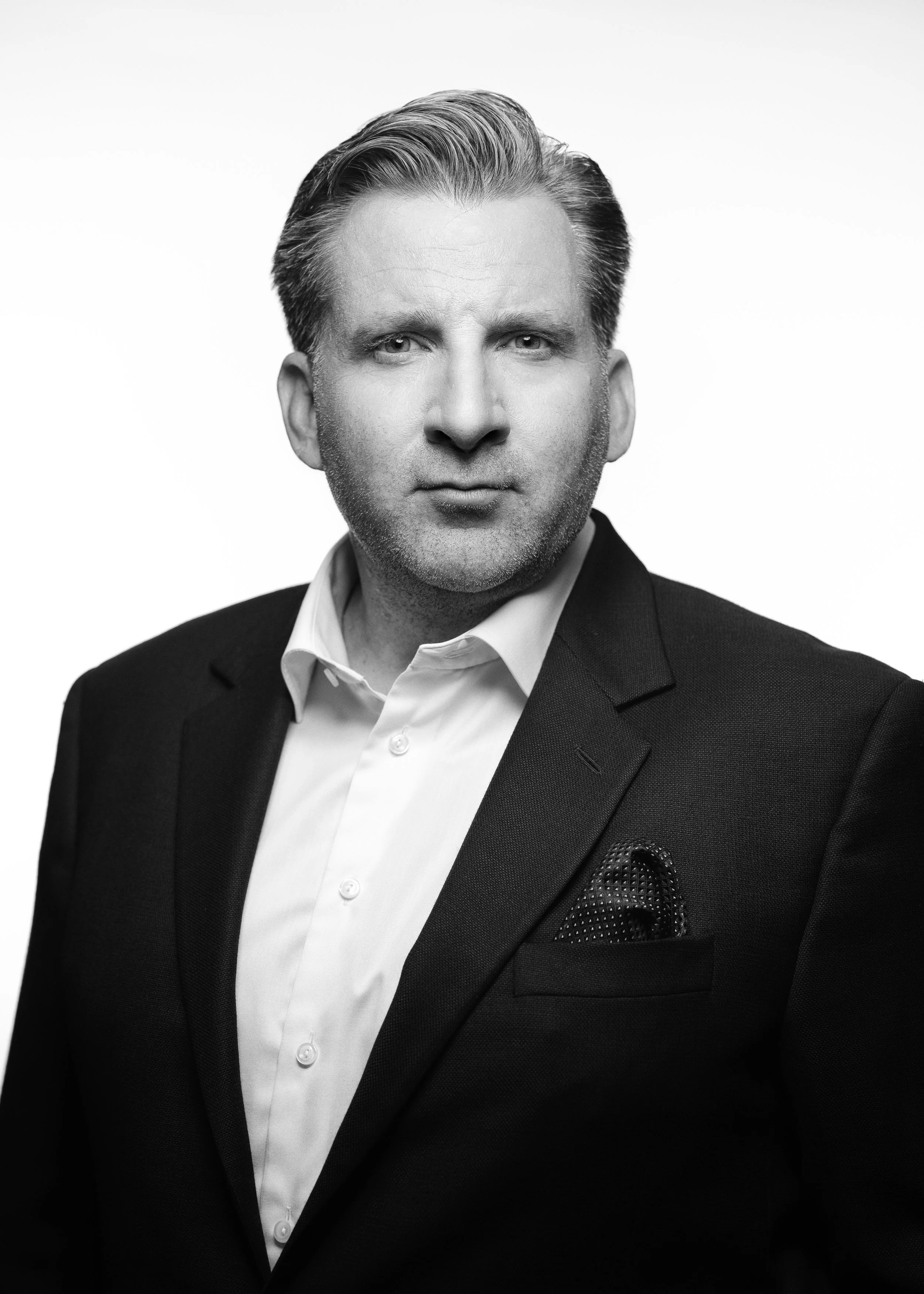 Black and white portrait of a man with neatly groomed hair, wearing a dark suit, white shirt, and patterned pocket square, against a plain light background.