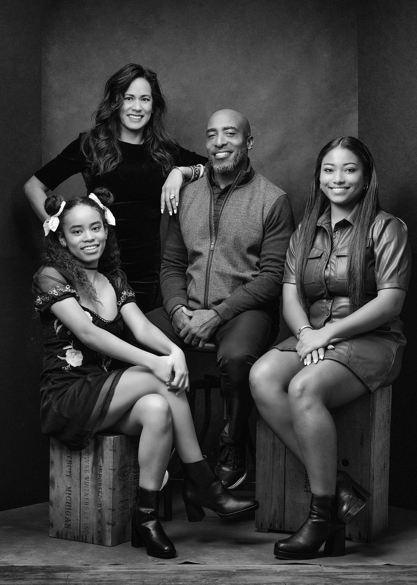 Black and white photo of a family of five posing together in a studio. The woman on the left has long wavy hair and is wearing a black dress. The man in the middle has a bald head and beard, wearing a zip-up vest over a striped shirt. The woman on th