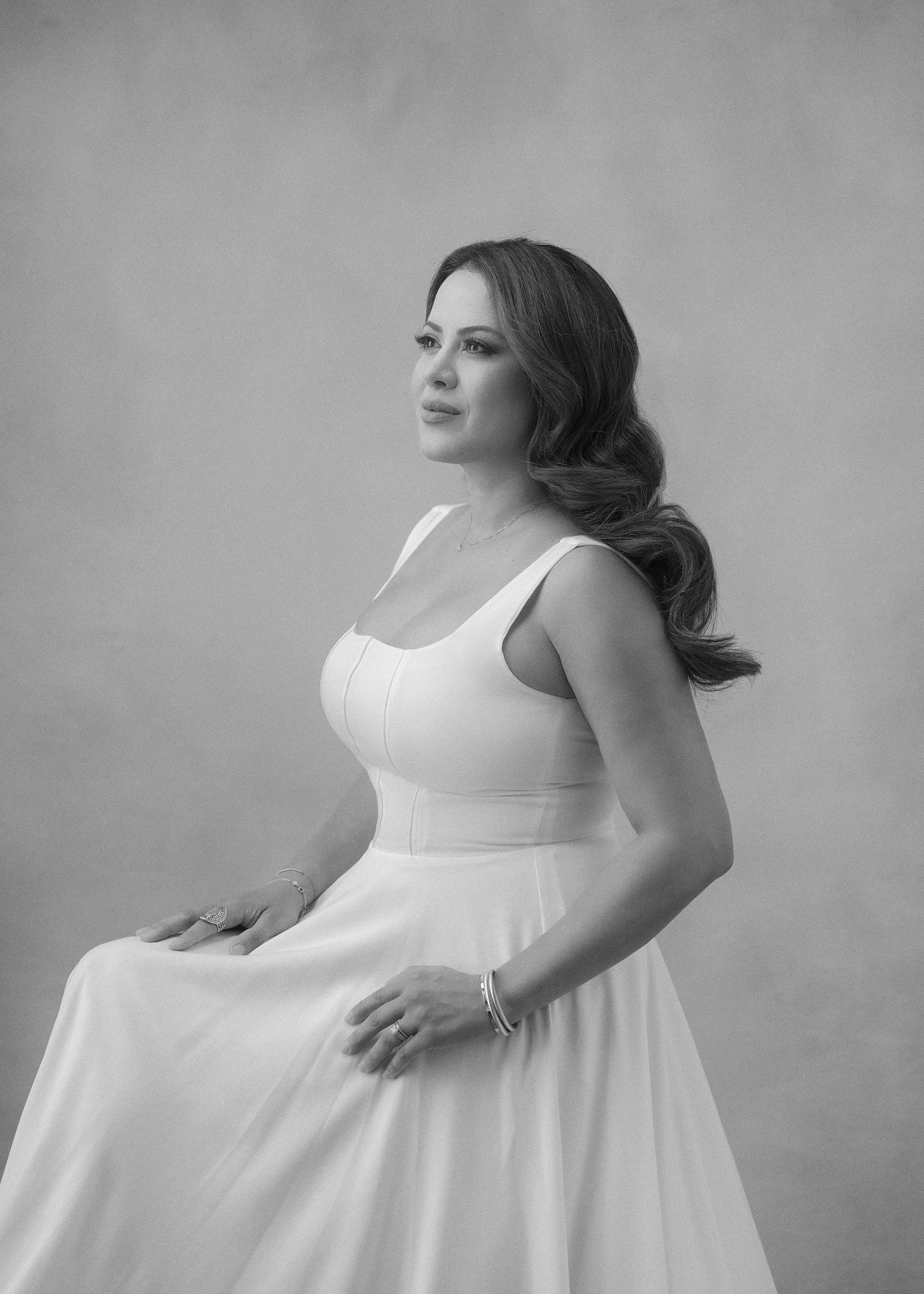 A woman with long wavy hair wearing a white gown sitting on a chair against a plain backdrop.