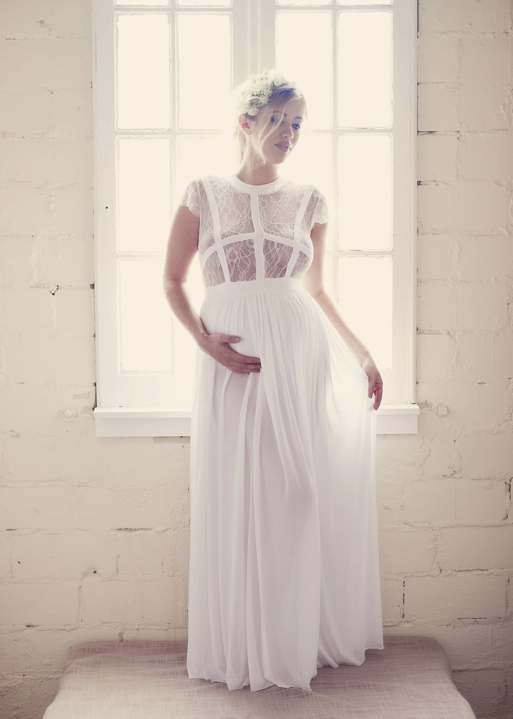 A woman standing on a bed in front of a window, wearing a white lace dress with cut-out details and a flower headpiece, with her hand resting on her pregnant belly.
