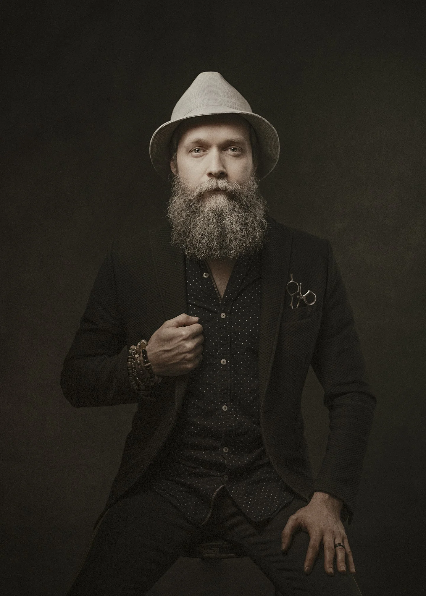 A man with a full beard and mustache wearing a white fedora, black suit jacket, and a dark shirt with polka dots, sitting against a dark background. He has a pair of scissors tucked into his jacket pocket and several bracelets on his left wrist. His 