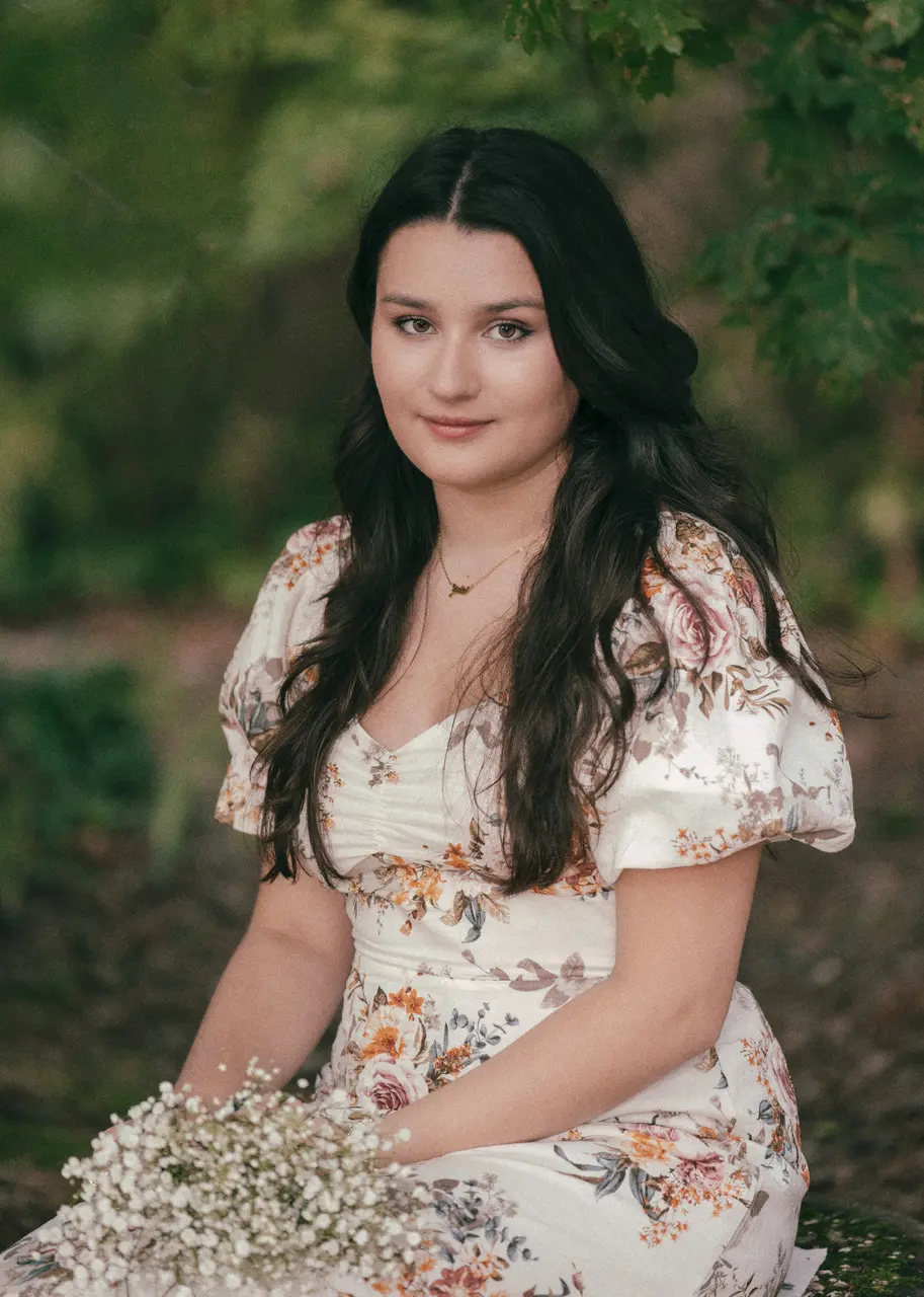 A young woman with long black hair wearing a floral dress sitting outdoors, holding a bouquet of white flowers, with trees and green foliage in the background.