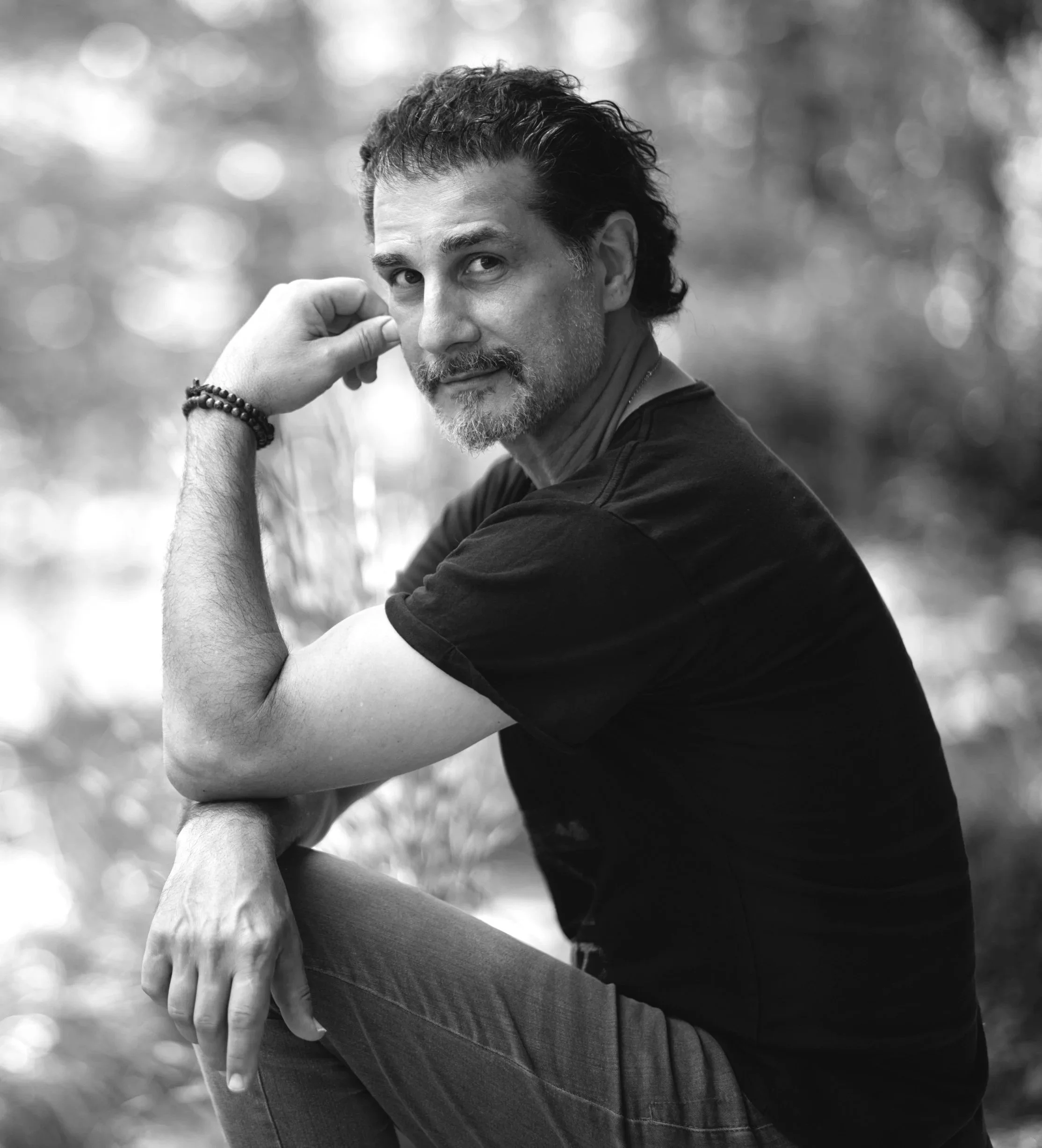 Black and white photo of a man with wavy hair and a beard, resting his face on his hand, outdoors with a blurred natural background.