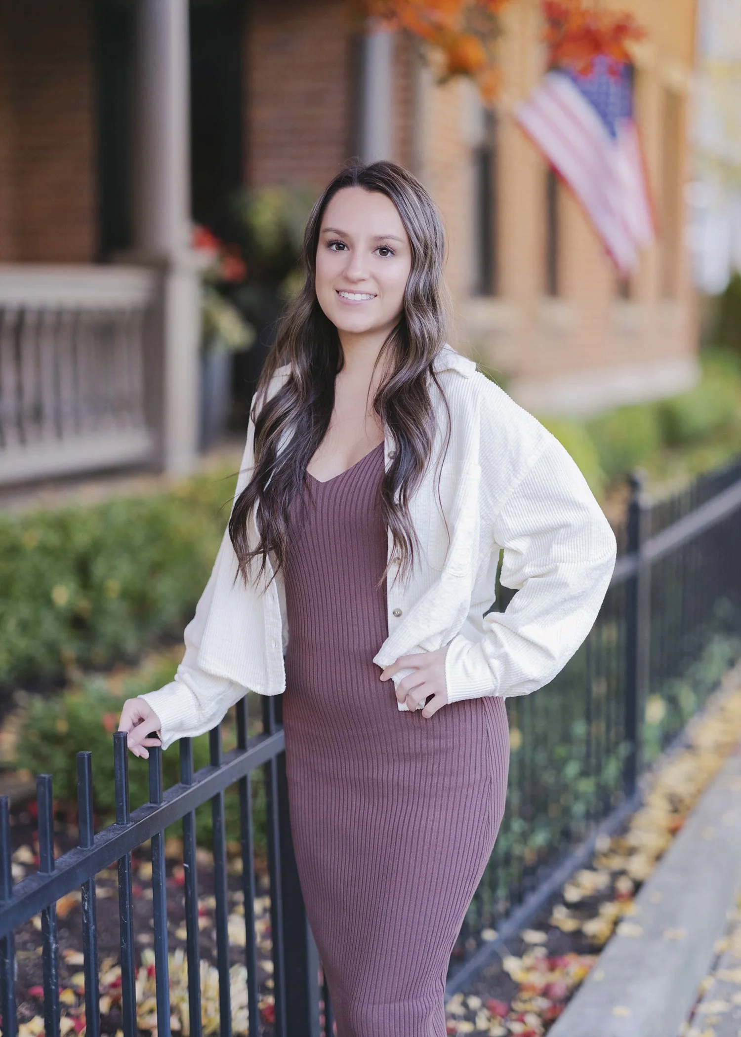 A young woman with long wavy dark hair, smiling, wearing a burgundy ribbed dress and cream-colored jacket, standing outdoors near a black metal fence, with fall-colored leaves and an American flag in the background.
