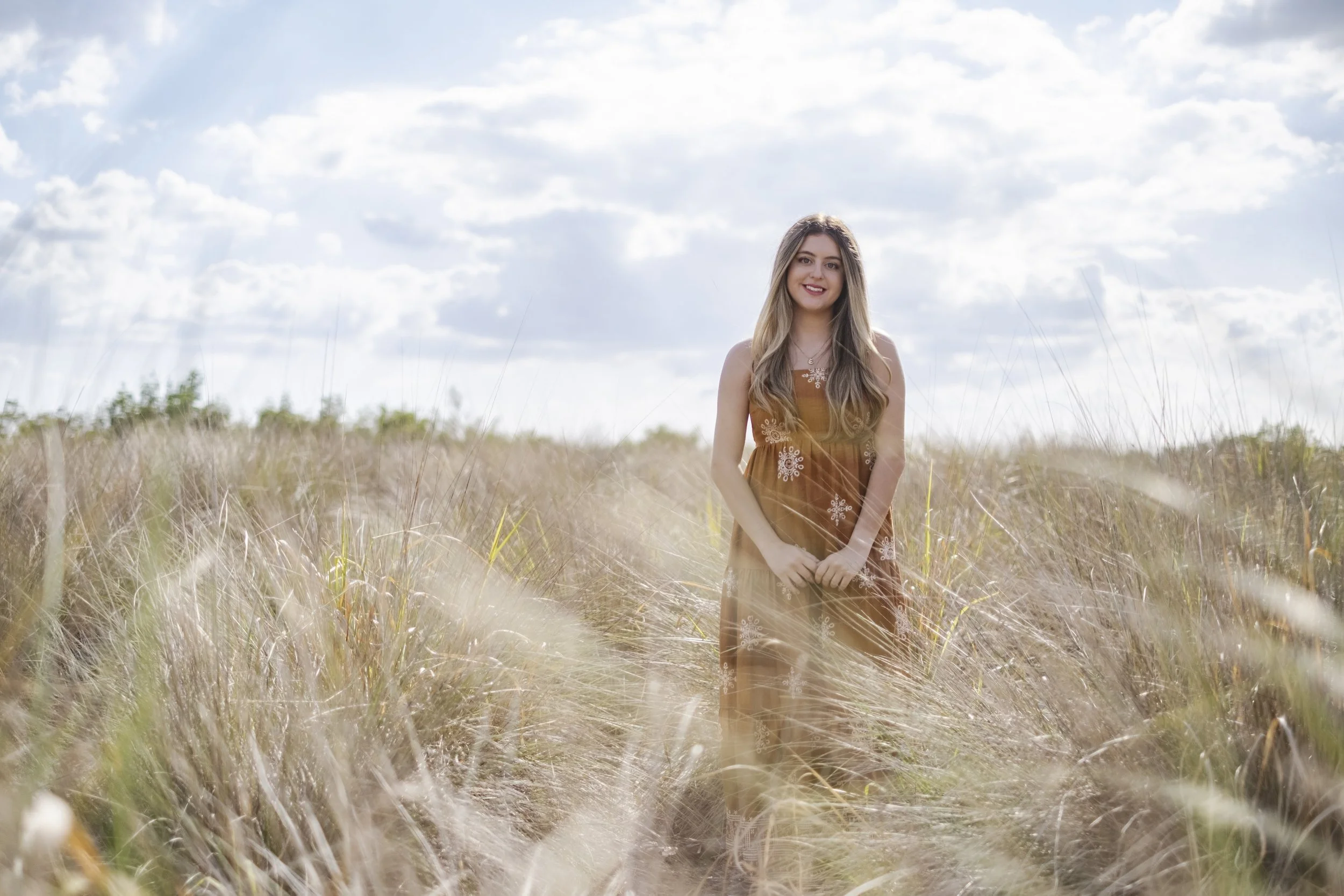 A young woman with long wavy hair wearing a brown dress standing in a field of tall grass under a partly cloudy sky, smiling at the camera.